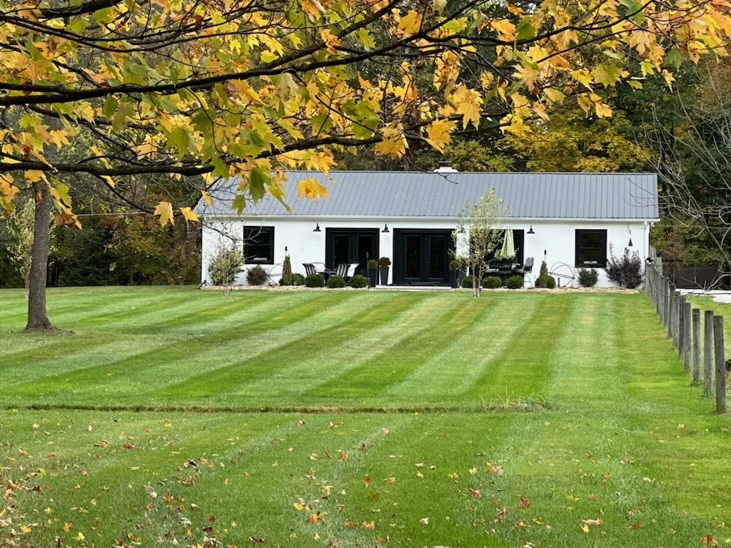 A white house with dark doors and windows in the background, a well-maintained green lawn with striped patterns, trees with changing autumn leaves, and a fence on the right side.