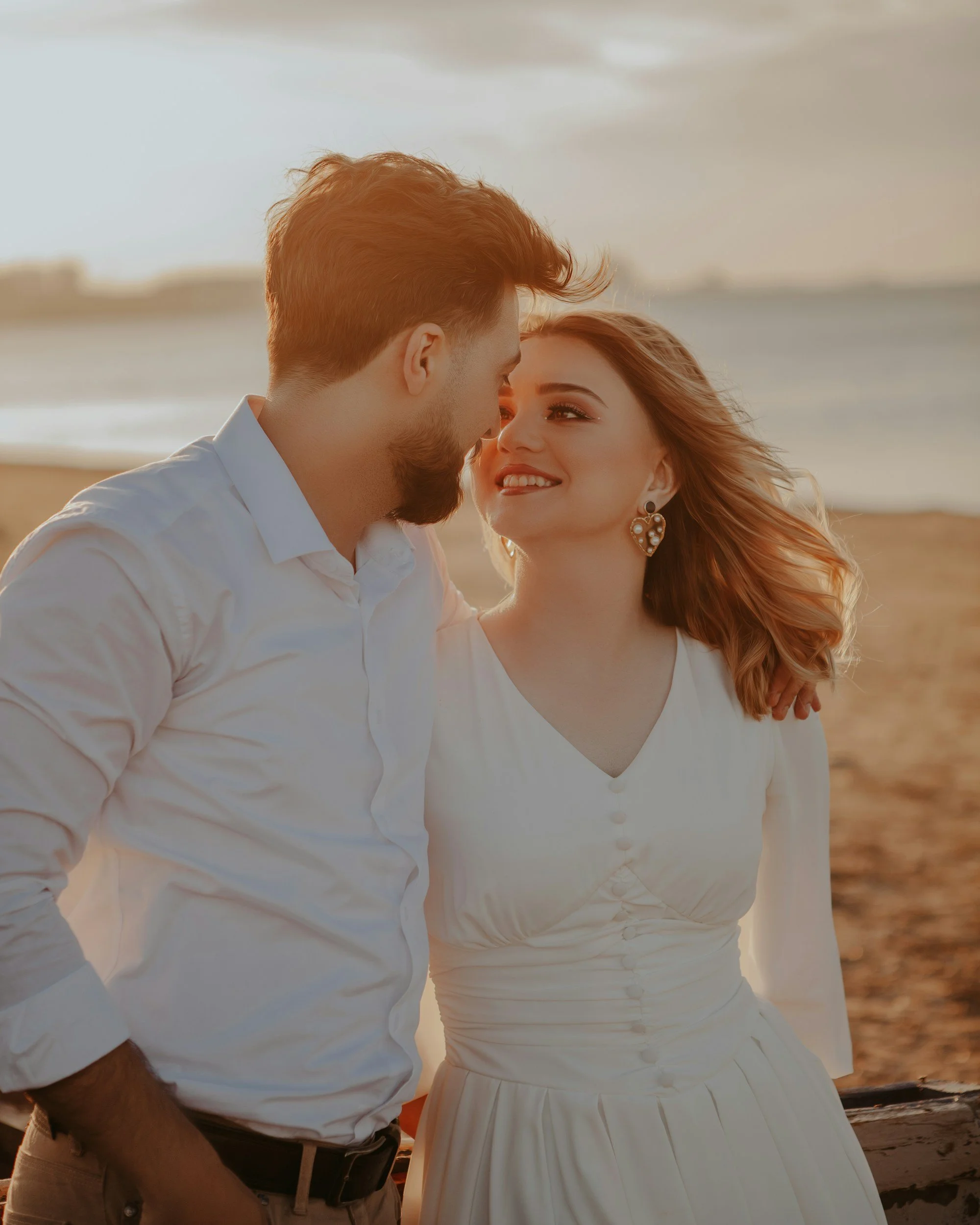 A close-up photo of a couple on the beach during sunset, smiling and looking at each other.