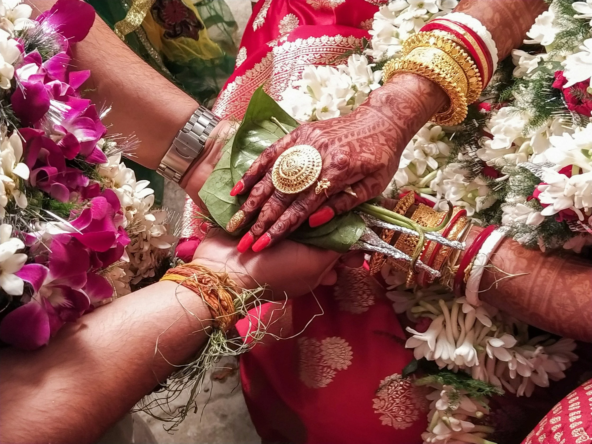 Handmade floral garland with pink, white, and purple flowers and green leaves, decorated with traditional gold jewelry and red fabric with gold embroidery, on a person's arm and hand covered with henna designs, wearing gold bangles and rings.