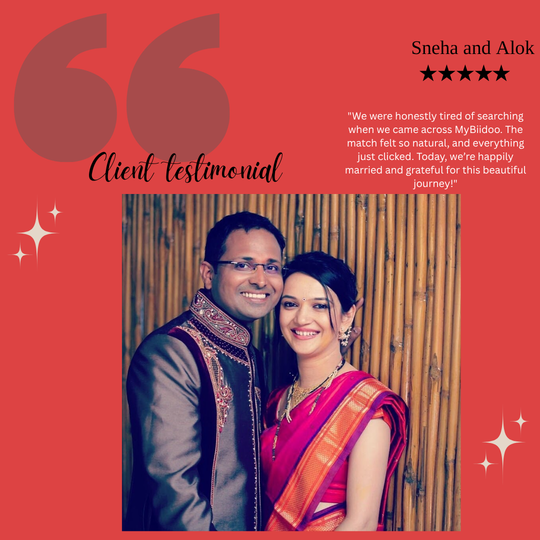 A man and woman dressed in traditional Indian attire, smiling and standing closely with bamboo background. The man is wearing a dark-colored sherwani with intricate embroidery, and the woman is wearing a pink saree with red and gold accents and jewelry.