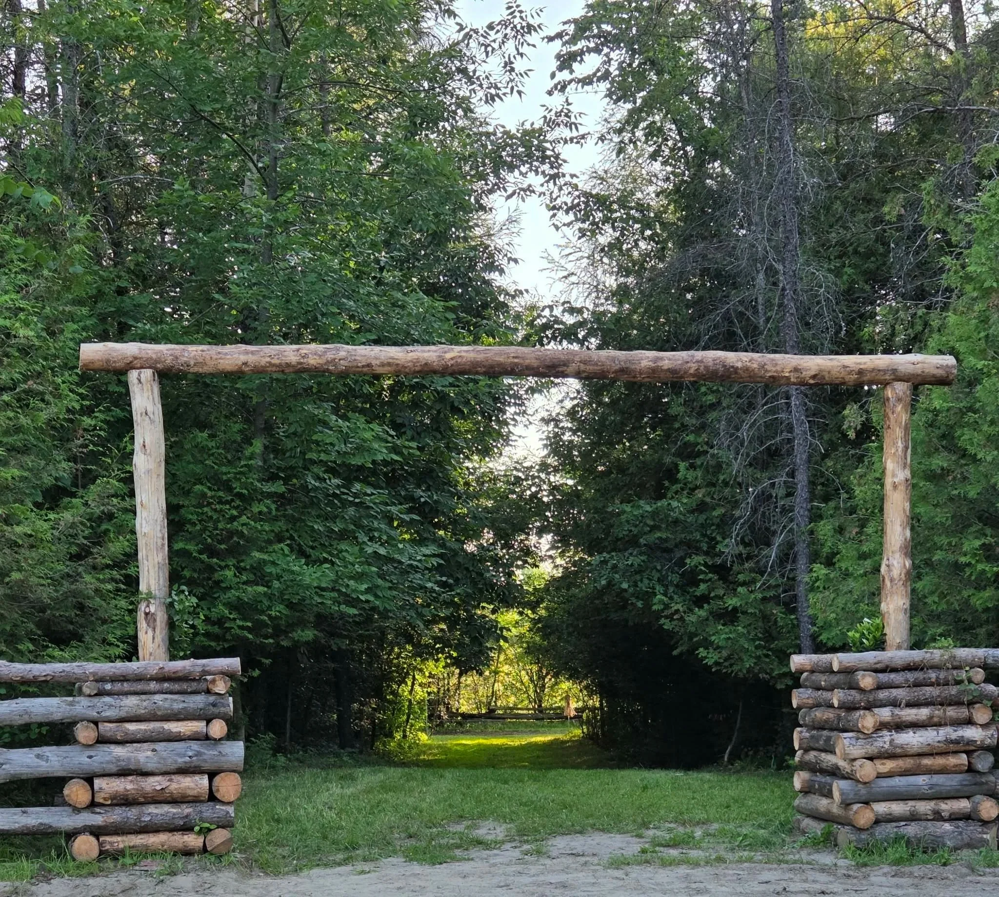 A tall arch made of logs comes out of a log fence and leads to a tree-covered lane and greenery.