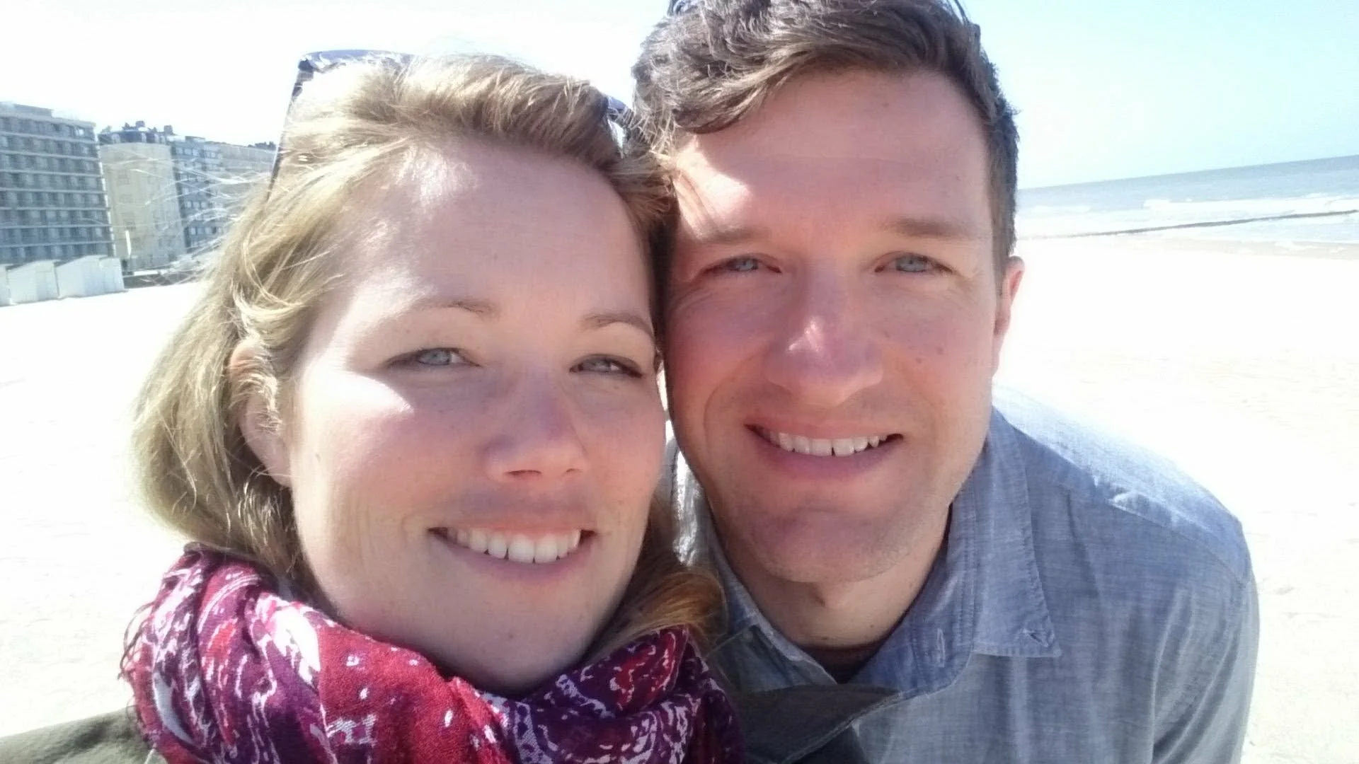 A smiling woman with light hair and blue eyes taking a selfie on a bright beach, standing next to a smiling man with light brown hair and blue eyes, with the ocean and beach in the background.