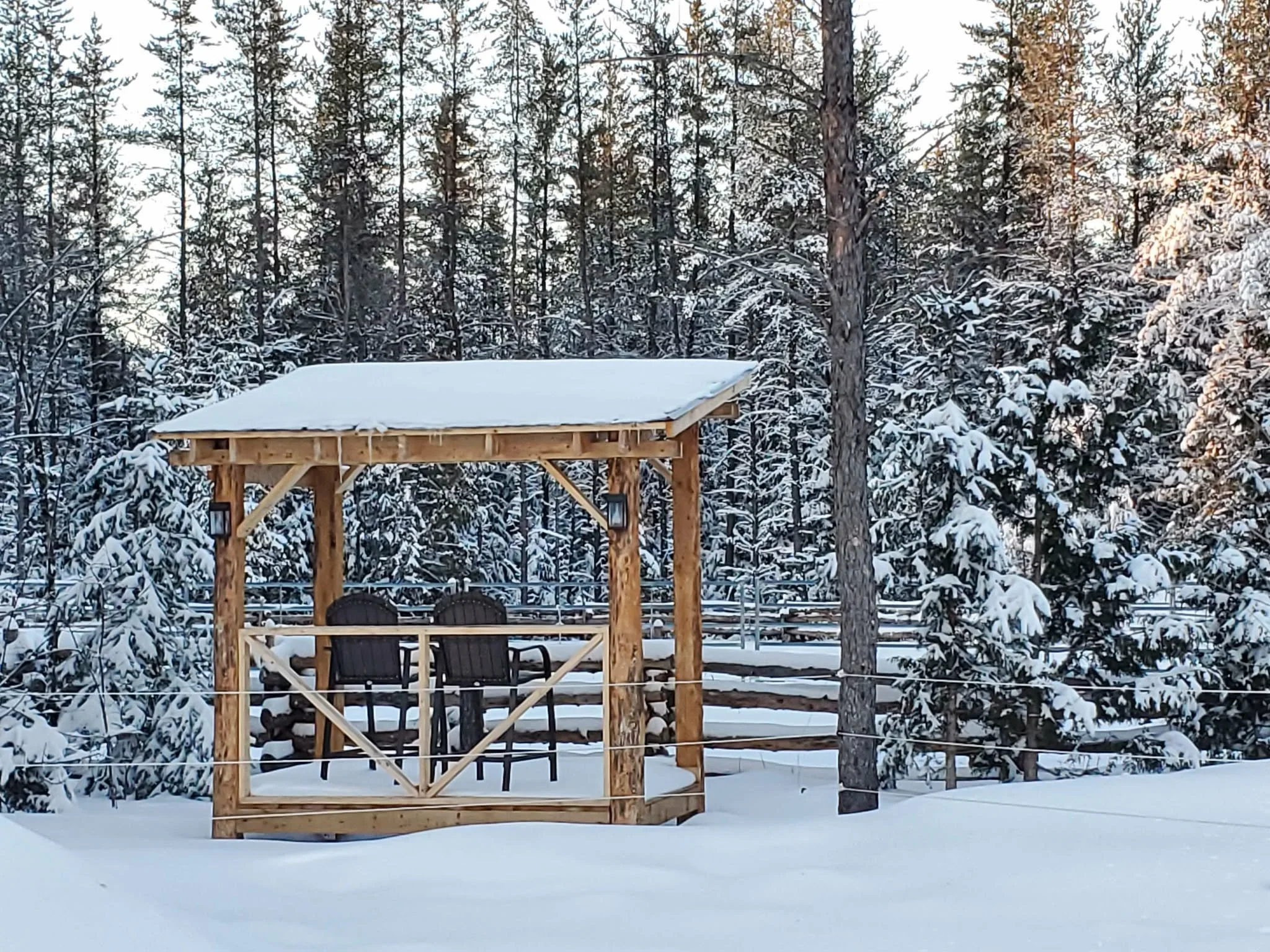 A snowy outdoor scene with a wooden gazebo and dark chairs, surrounded by snow-covered trees in a forest.
