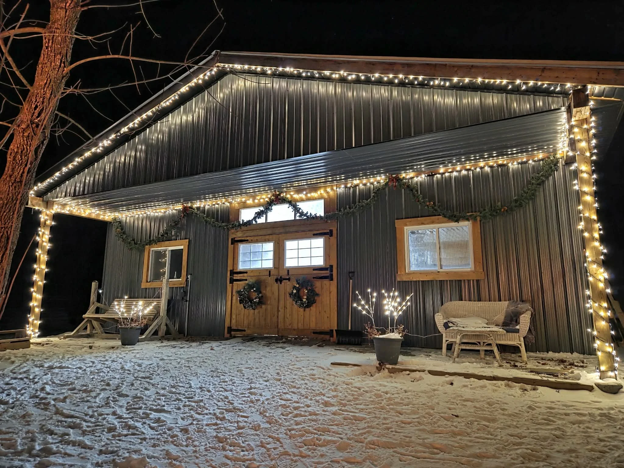 A nighttime celebratory or elopement scene of the exterior of a barn decorated with many Christmas lights. There is a wooden door with holiday wreaths and the yard is covered with snow.