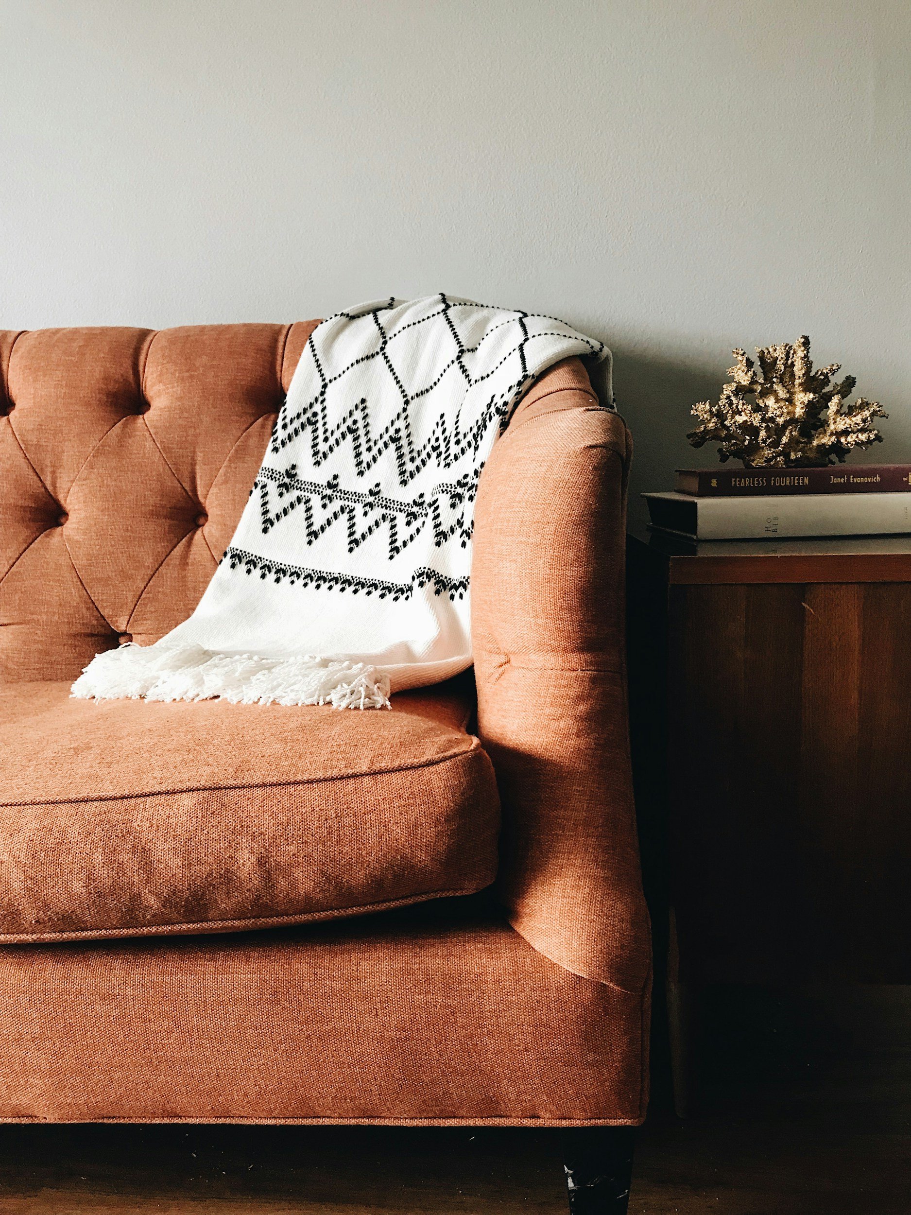 A burnt orange tufted sofa with a black and white patterned throw blanket draped over one arm. Next to the sofa is a dark wooden side table with a small dried flower arrangement and some books.