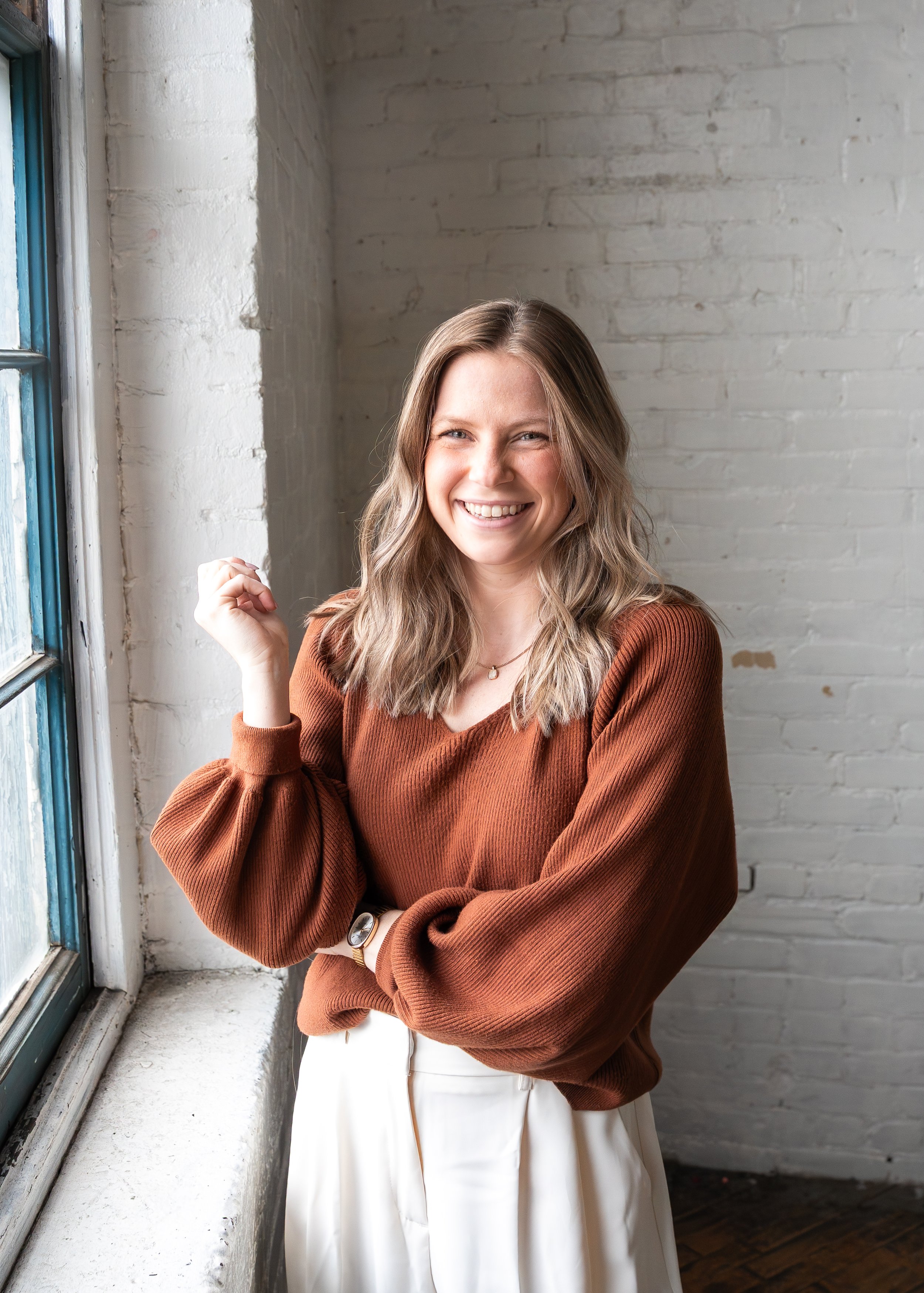 A woman with long, light brown wavy hair, smiling, standing indoors near a large window with a white brick wall in the background. She is wearing a rust-colored long-sleeve top, white pants, and a watch on her left wrist.