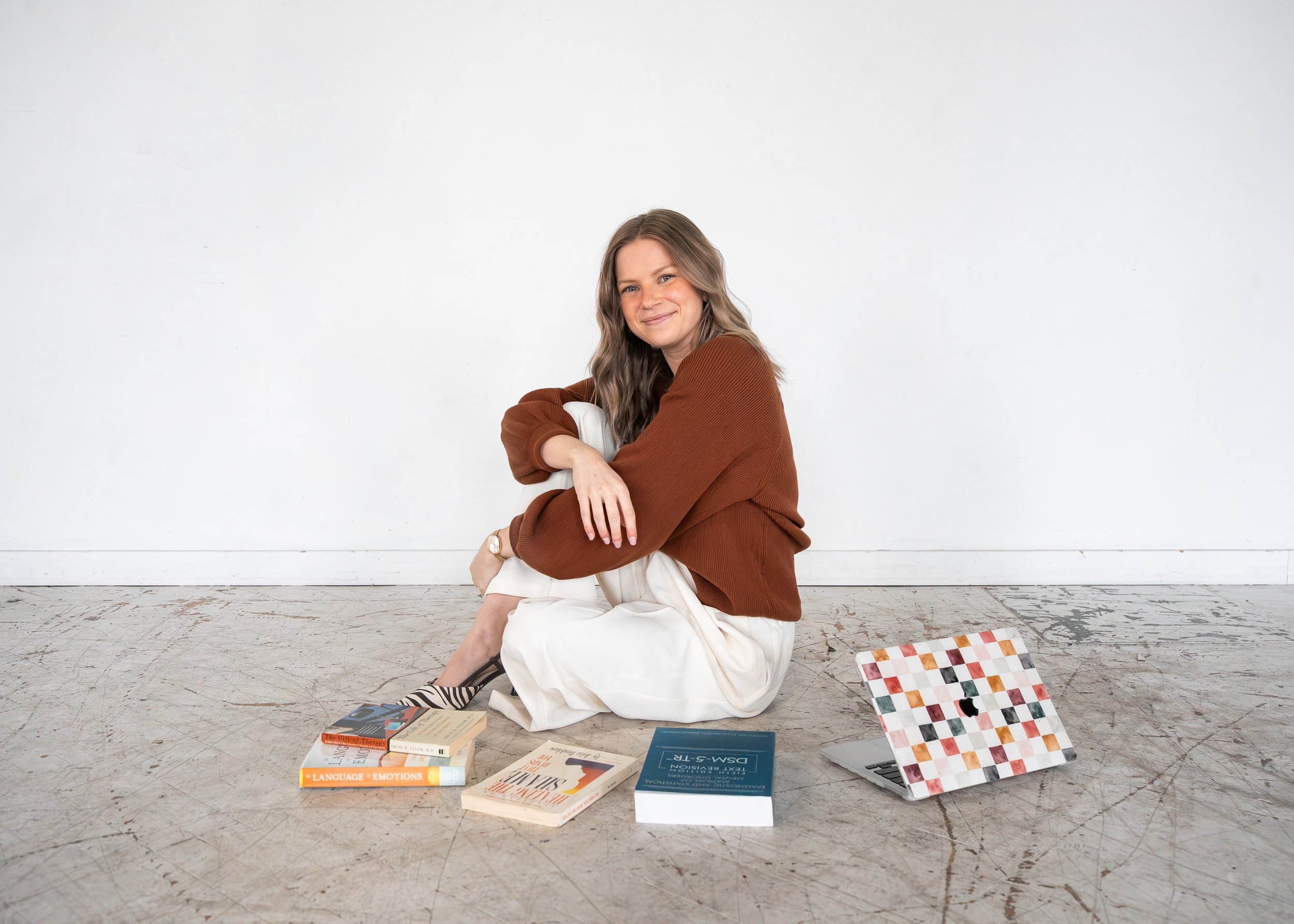 A woman with wavy brown hair, smiling, sitting on the floor against a white wall, surrounded by books and a color palette board, wearing a brown sweater and beige pants.