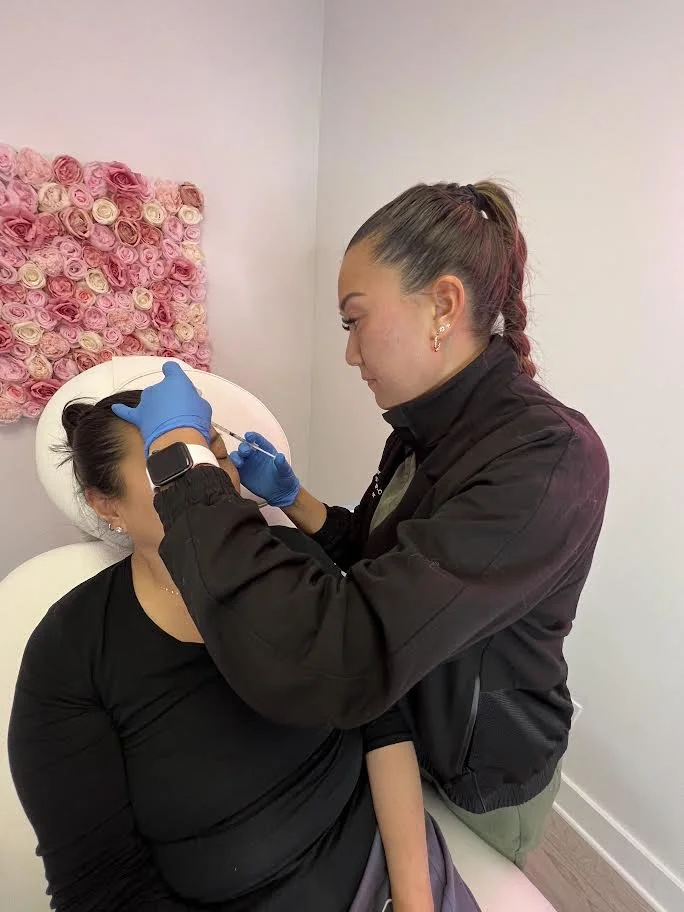 A woman with gray hair tied in a ponytail receiving cosmetic injections from a healthcare professional wearing blue gloves, in a clinic with a floral wall decoration in the background.