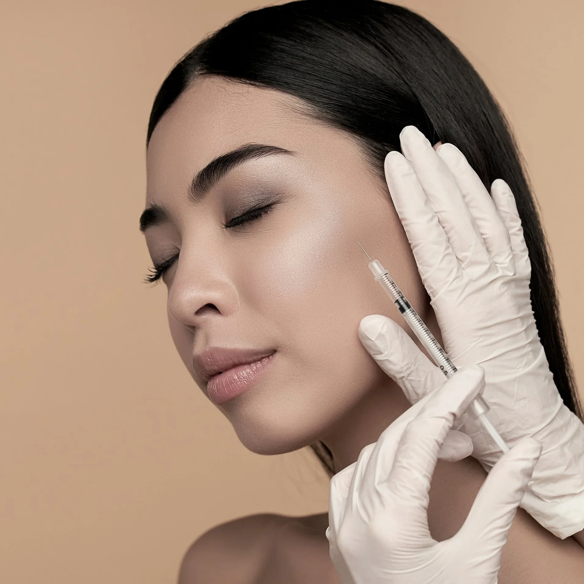 A woman with closed eyes and flawless skin receiving a cosmetic injection on her cheek from a medical professional wearing white gloves.