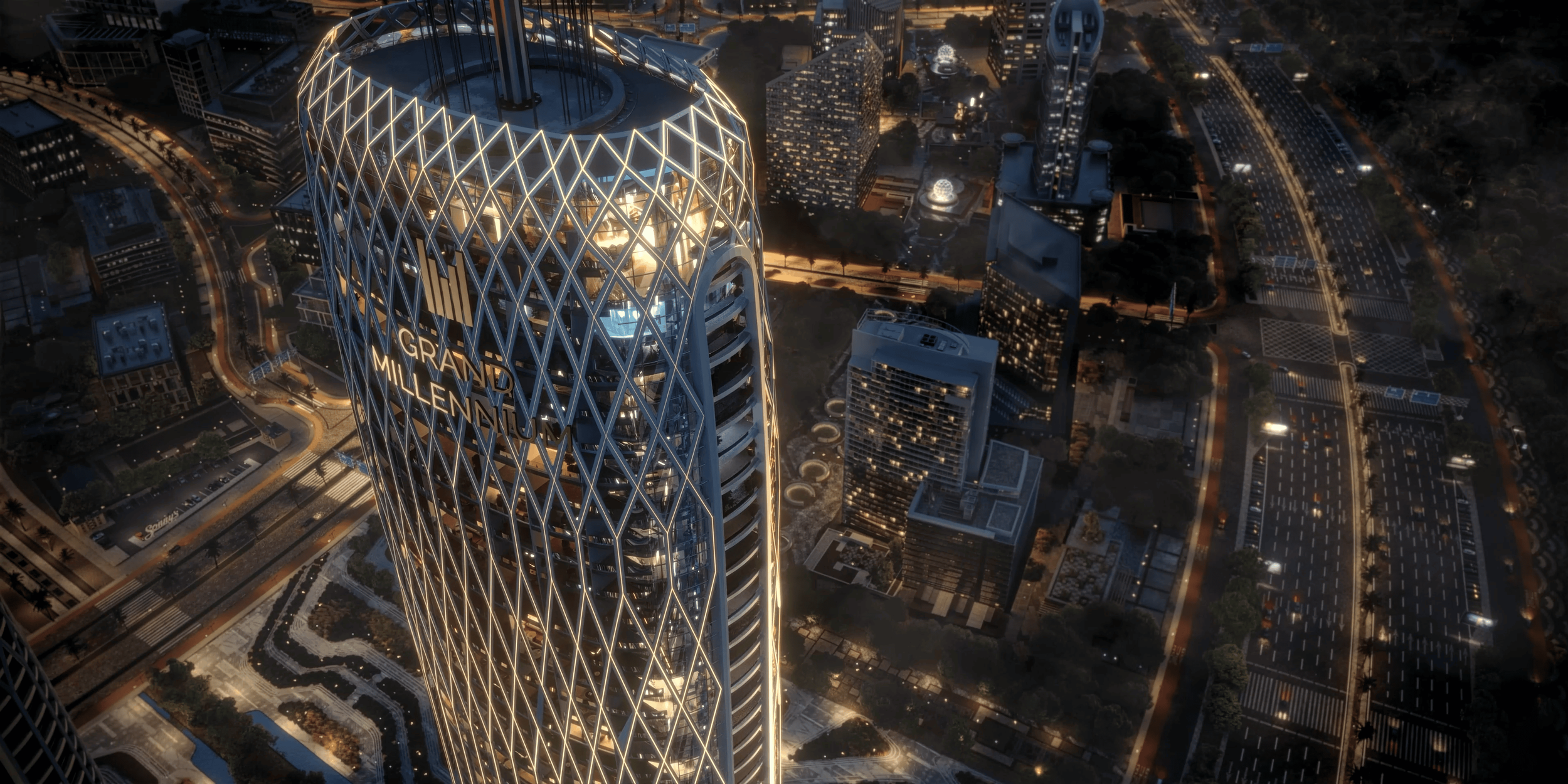 Night aerial view of a modern city with a tall skyscraper labeled 'Grand Millennium' illuminated by lights, surrounded by other buildings, roads, and green spaces.