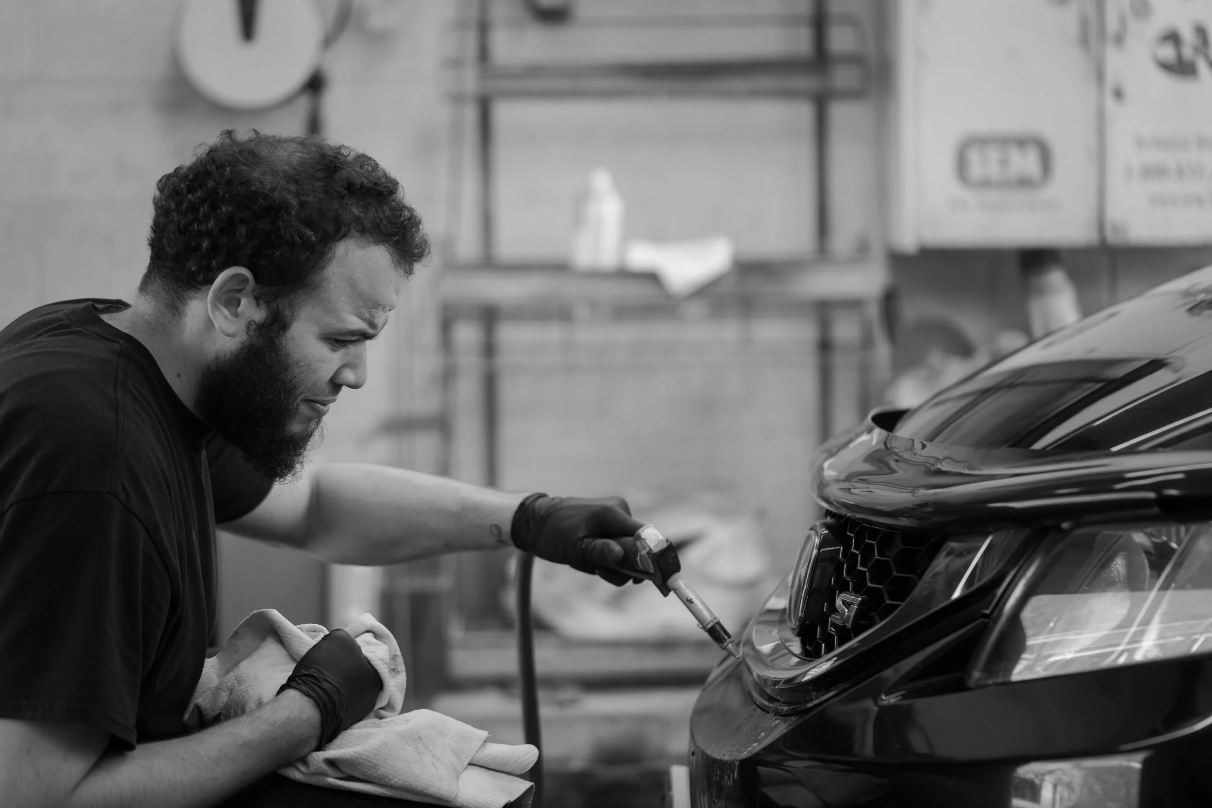 A man with curly hair and a beard is polishing or detailing a car's front grille with a cloth in an auto shop or garage.