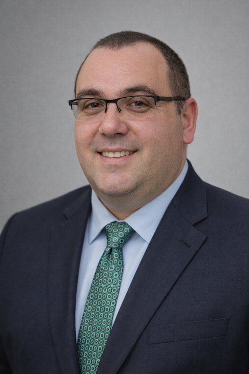 Portrait of a man wearing glasses, a navy blazer, light blue shirt, and patterned green tie, smiling against a gray background.