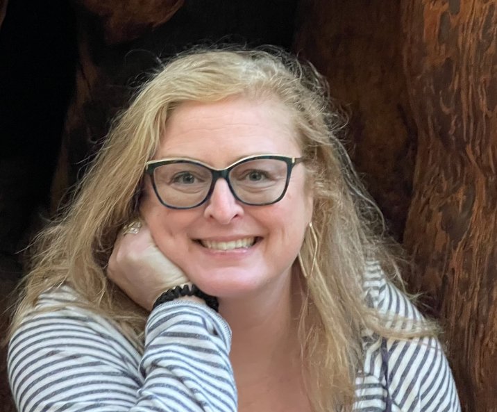 Close-up of a smiling woman with blonde, wavy hair, wearing glasses and a striped shirt, seated indoors with a wooden background.