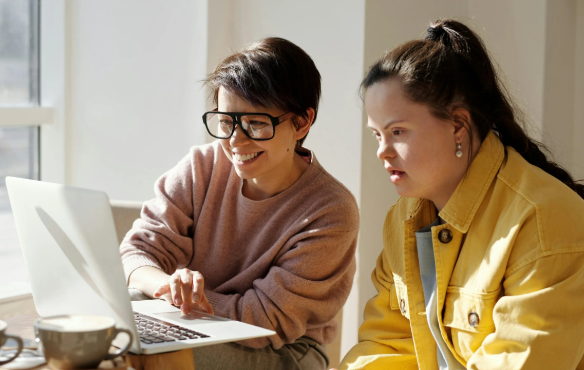 Two women sitting at a table looking at a laptop, one is smiling and wearing glasses, the other appears focused, with cups on the table, in a bright room.