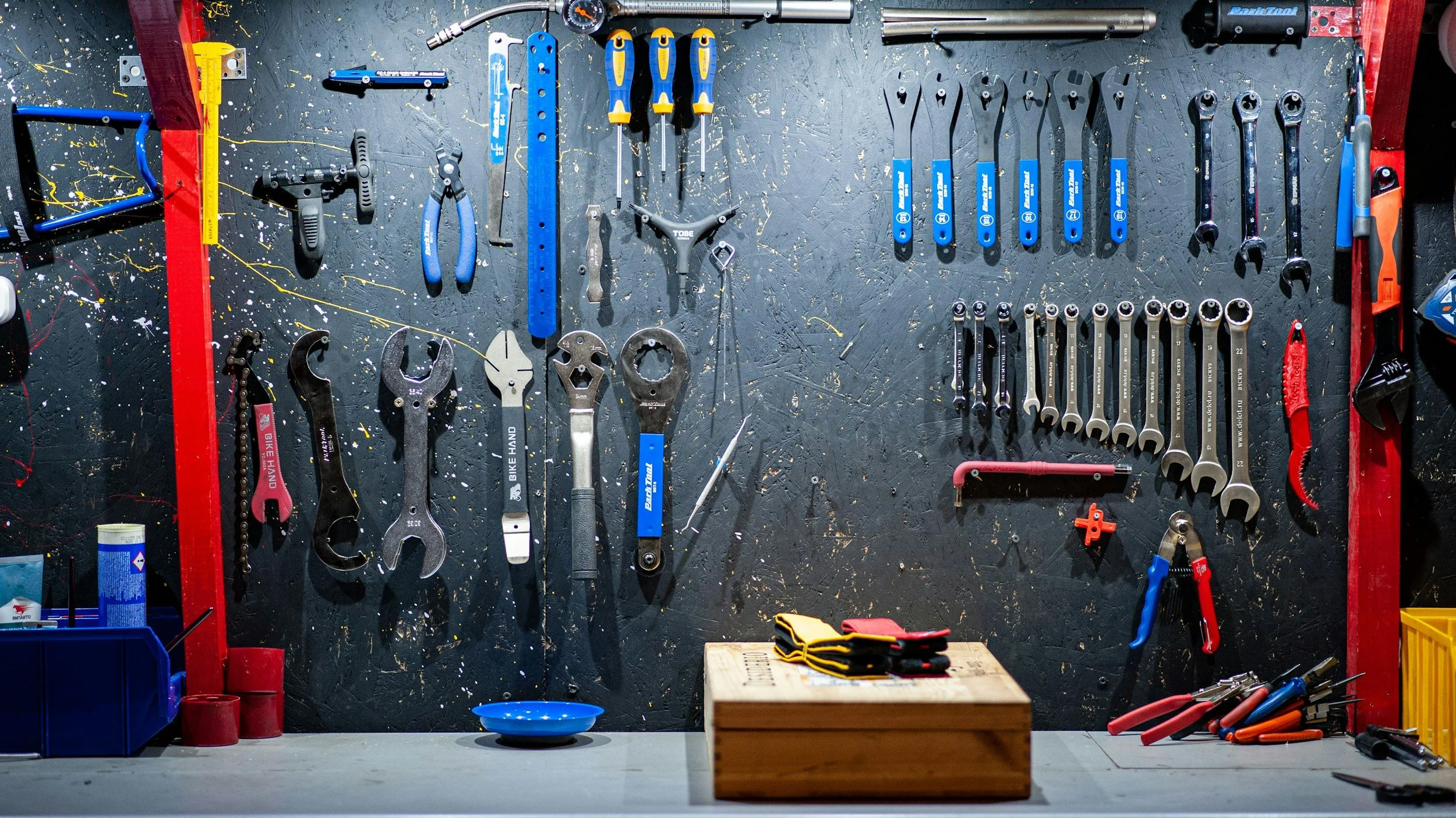 A black pegboard wall holding various hand tools including wrenches, screwdrivers, and pliers, with a workbench and yellow plastic crate below.