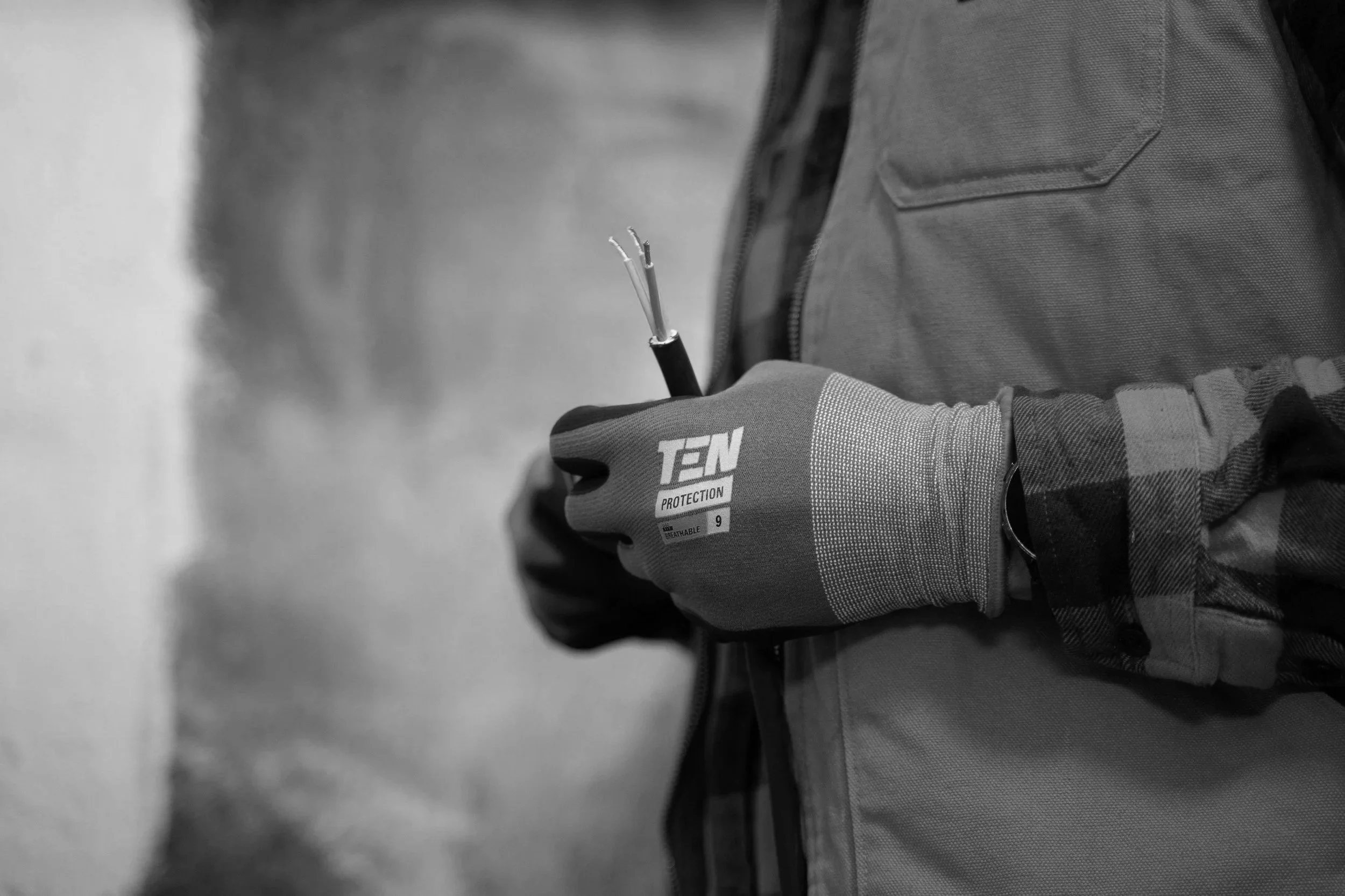 A person wearing a protective glove and a jacket, holding a pair of pliers with wires, in black and white.
