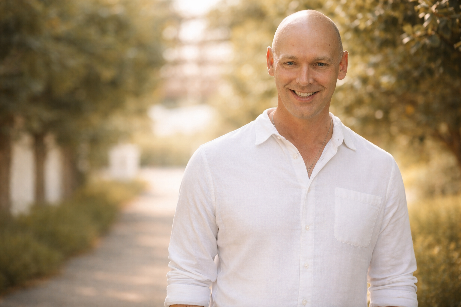 A smiling man with a shaved head, wearing a white button-up shirt, standing outdoors on a path lined with trees in autumn.