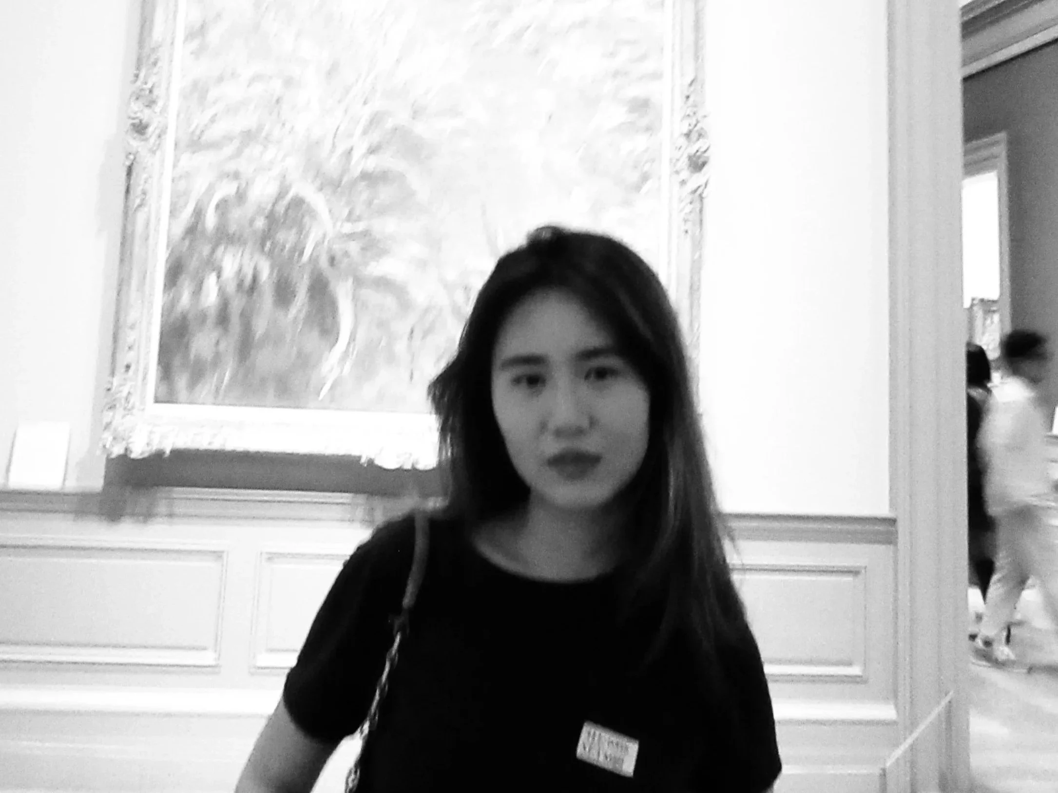 A young woman with long dark hair standing indoors, with a large painting behind her and a few other people blurred in the background.