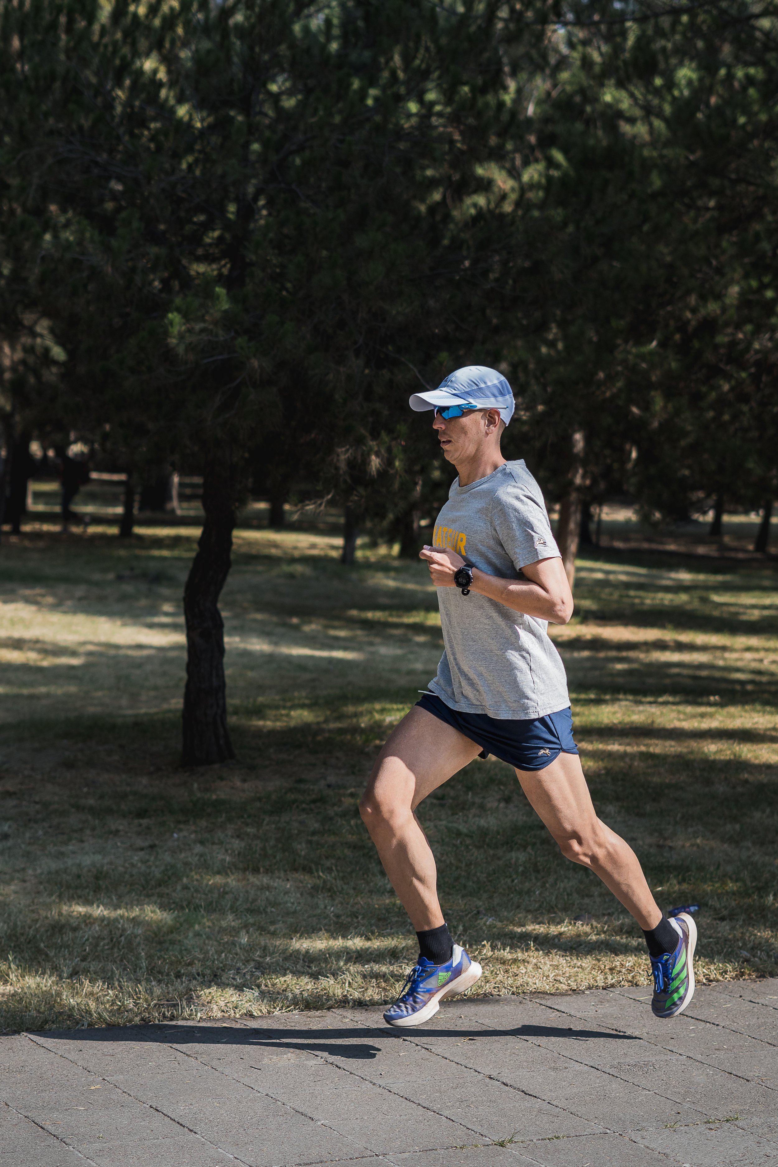 Un hombre corriendo por un parque en un día soleado, viste camiseta gris, gafas deportivas y gorra, lleva reloj en la muñeca y usa pantalones cortos azules y zapatillas de correr azules y verdes.