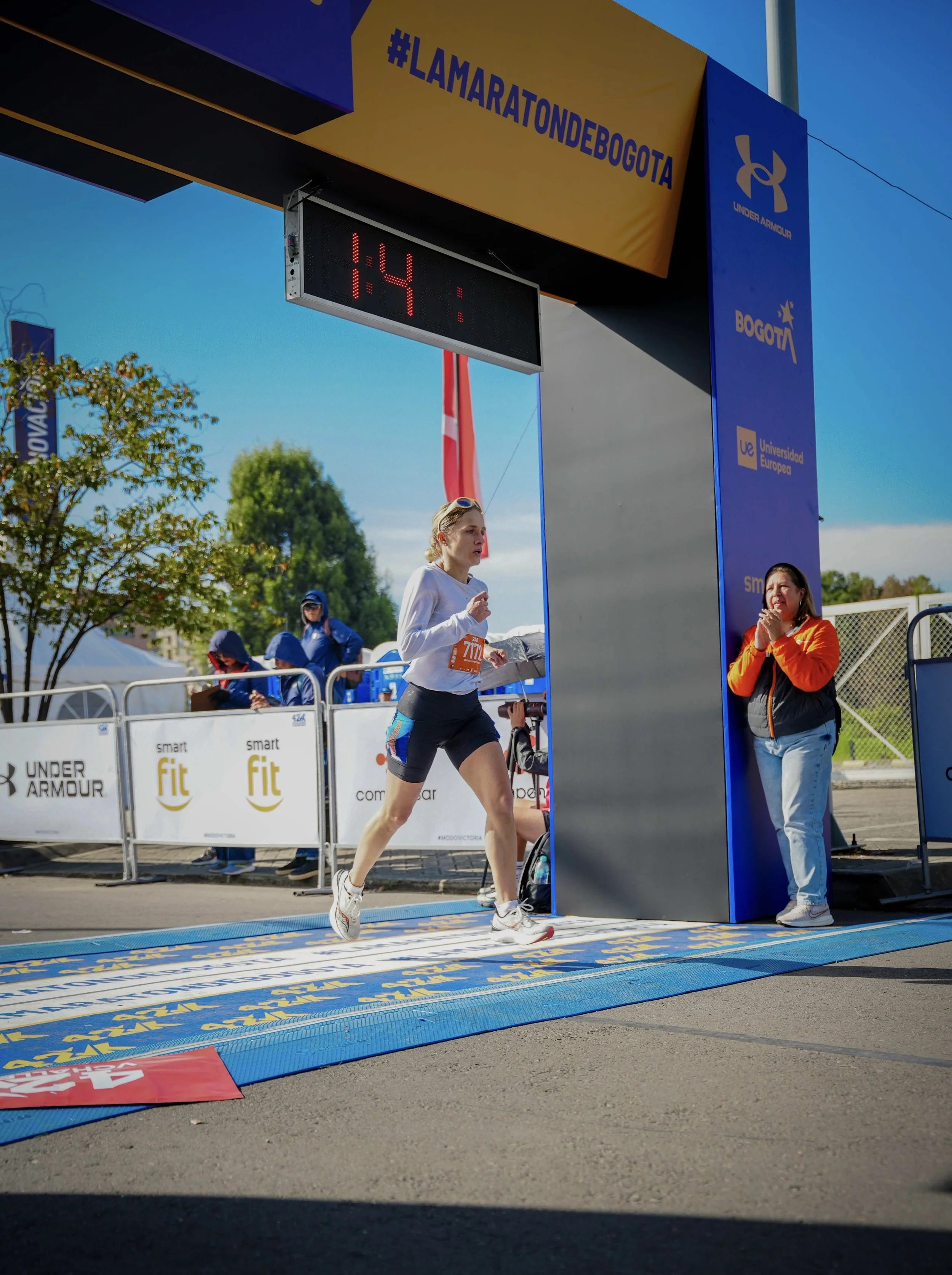 Mujer cruzando la línea de meta en una carrera en Bogotá, con un reloj digital que muestra 1:41, y un arco de meta azul con logos y anuncios. Visten ropa deportiva y llevan un número de participación.