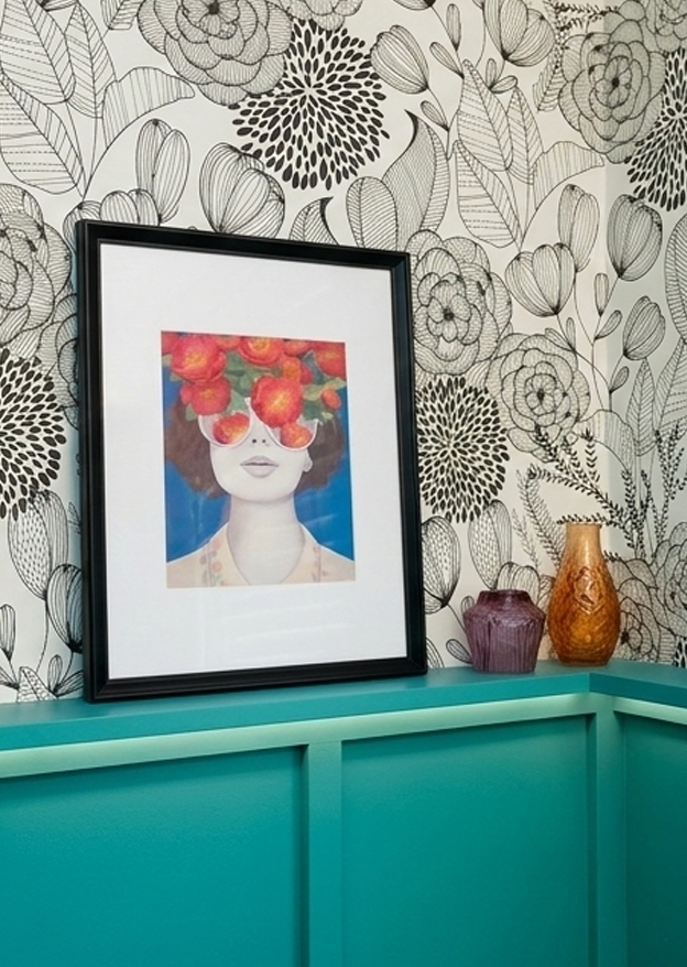 A framed digital art portrait of a woman with a head of orange and red flowers, set against a blue background, displayed on a teal shelf with two vases nearby, against a black and white floral patterned wallpaper.