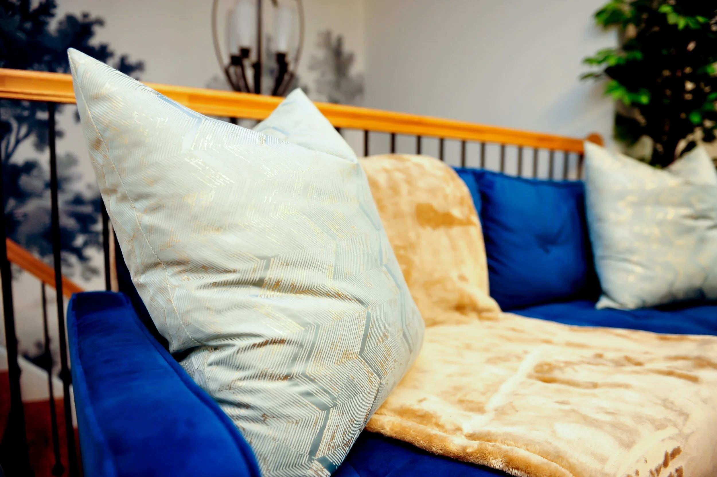 Close-up view of a loveseat with decorative pillows in various colors, including white, beige, and blue, against a headboard with a light wood frame and black metal bars in a bedroom with neutral walls, wall art, and a plant in the background.