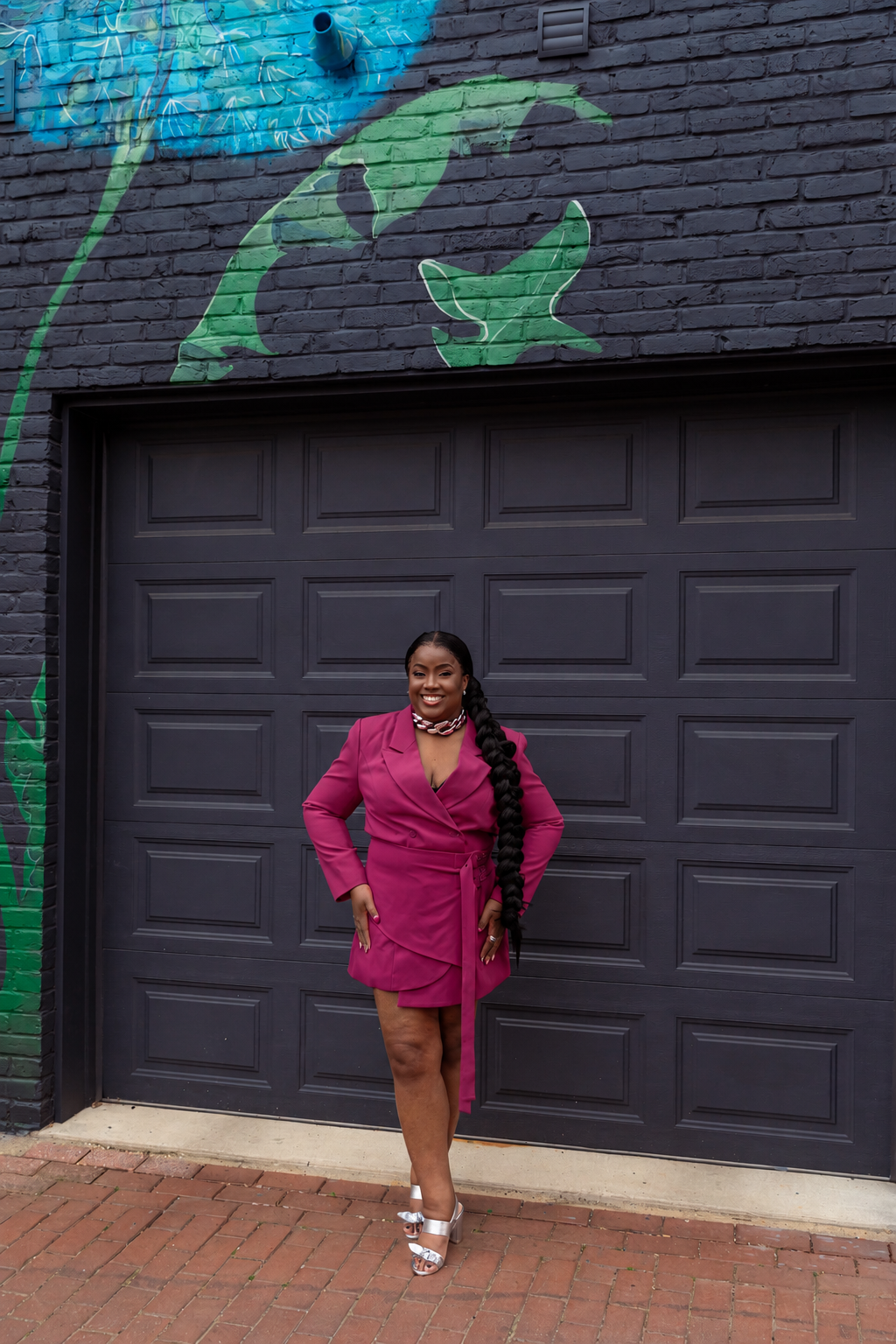 Pam standing in front of a dark gray garage door with a mural of a green fish and blue water painted above it. She is wearing a magenta dress, silver sandals, and has a long braid. She is smiling and posing with one hand on her hip.