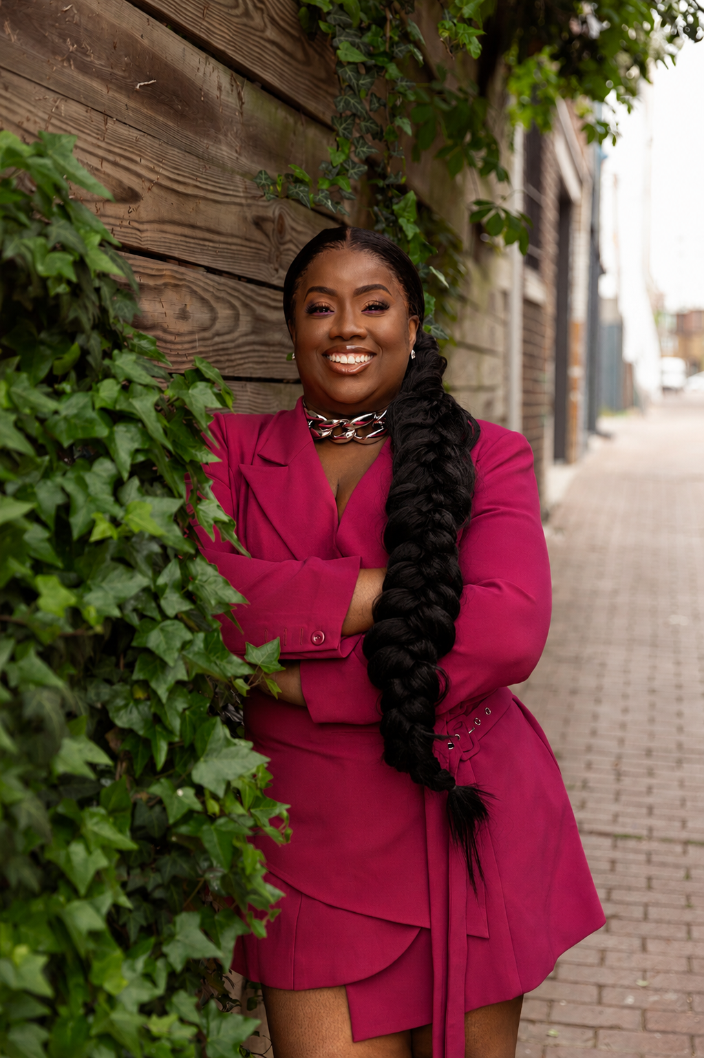 Pamela Agava, owner and smiling woman with long black hair in a thick braid, wearing a magenta blazer dress and a chunky silver necklace, standing outdoors next to a wooden fence covered with green ivy on a city sidewalk.