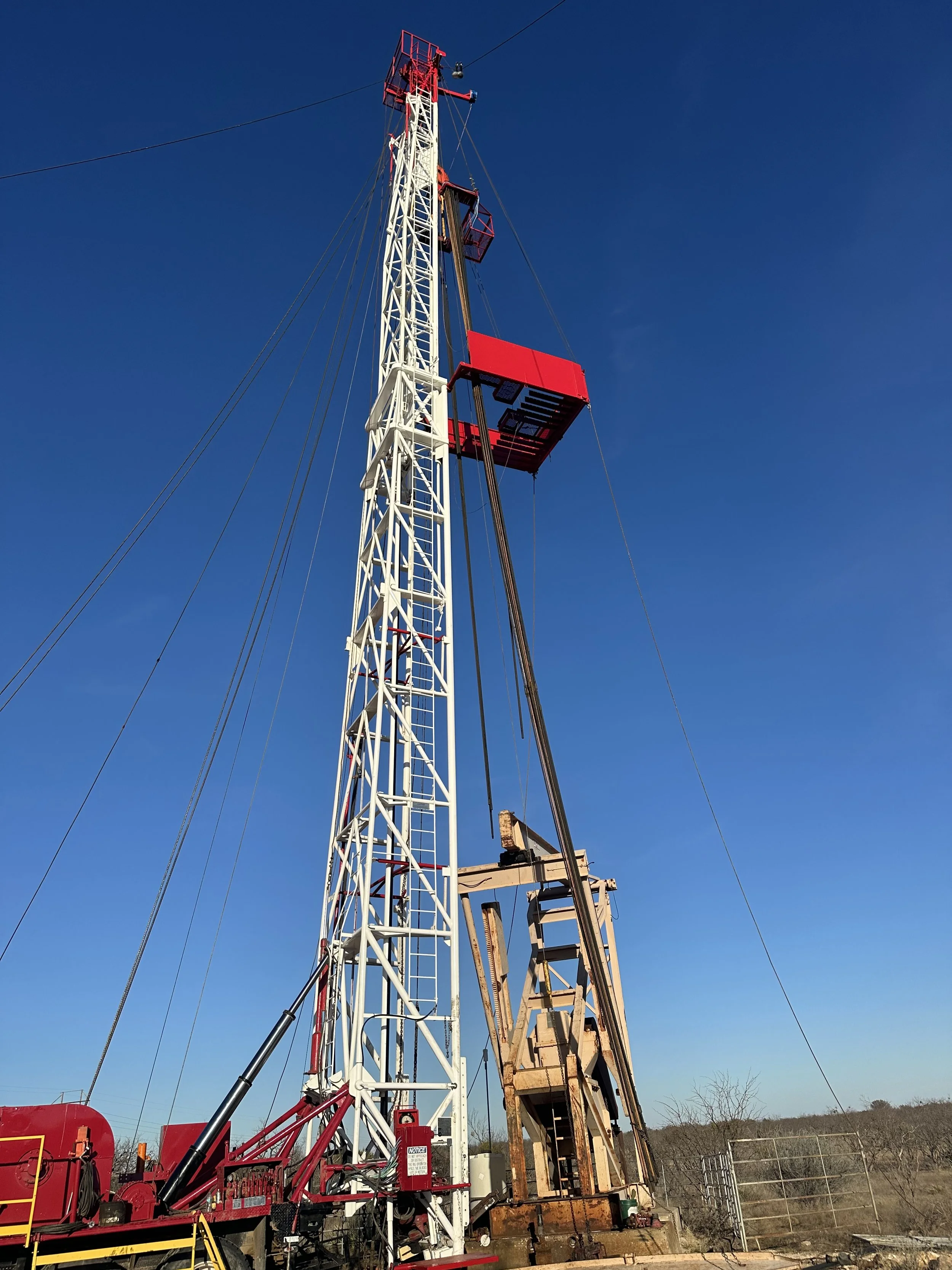 A tall oil drilling rig with a red and white structure, set against a clear blue sky, with an old rusted drilling platform nearby and some fencing in the background.