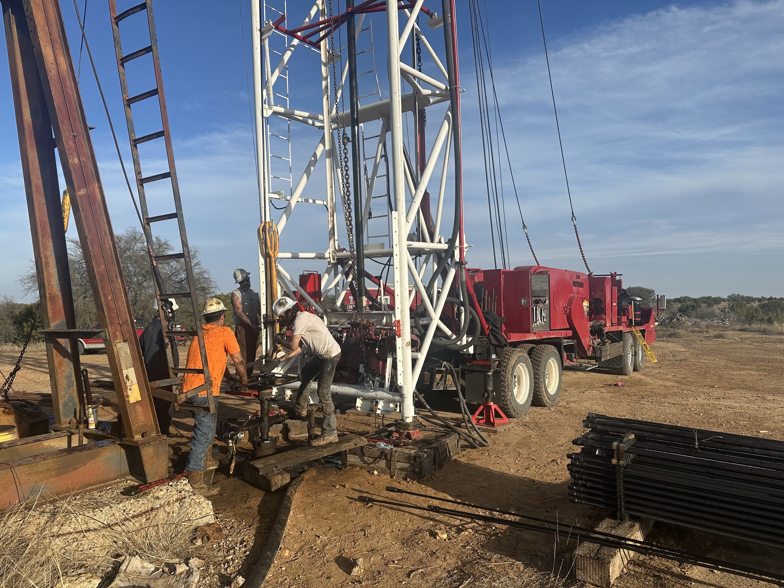 Workers in safety gear are assembling or maintaining a large red and white oil drilling rig on a dirt site, with a stack of black pipes nearby and a clear sky in the background.