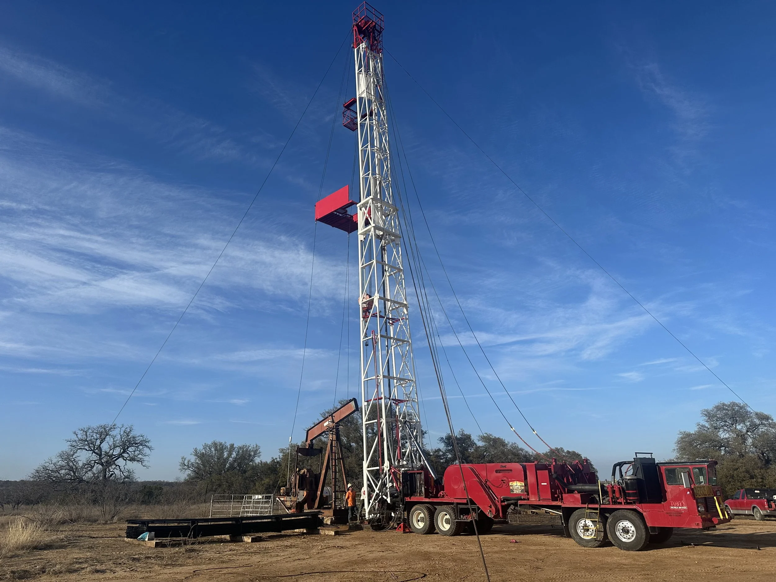 A tall oil well drilling rig in a rural area under a blue sky, with sparse trees and a red truck nearby.