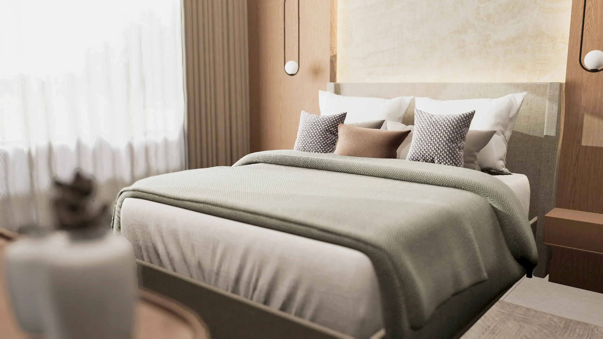 A neatly made bed with a beige headboard, white and patterned throw pillows, and a greenish-gray blanket in a modern hotel room with natural light from a window.