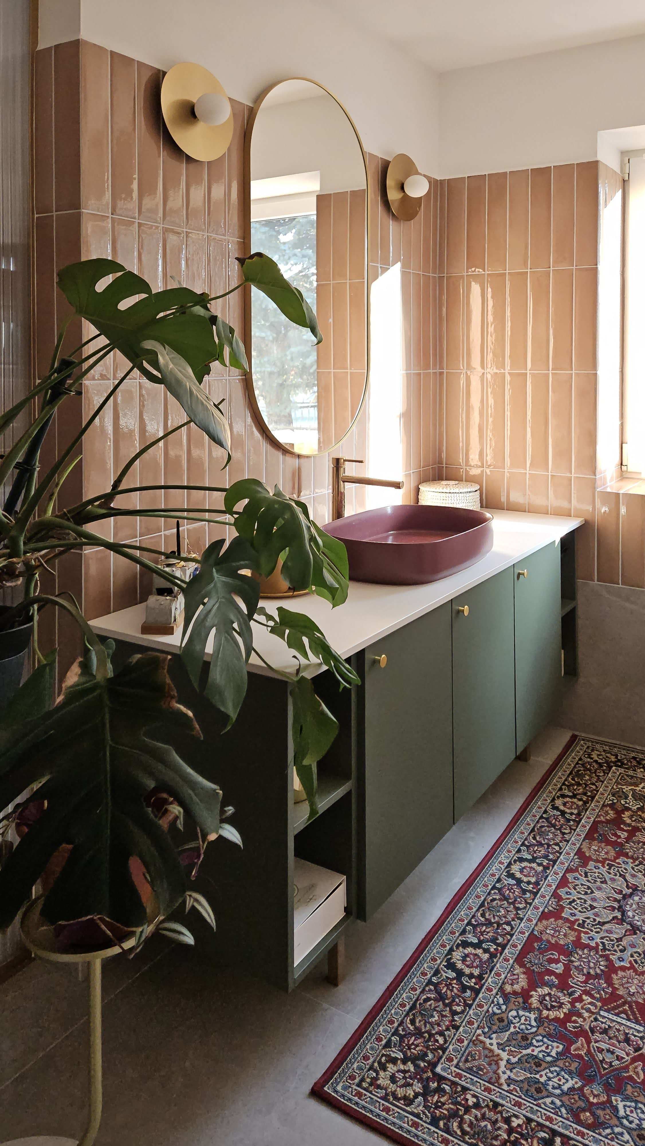 Bathroom with pink tiled walls, a green cabinet with gold knobs, a maroon sink, a tall mirror, two wall lights with gold accents, a window, a potted plant with large green leaves, a patterned rug, and a small wicker basket.