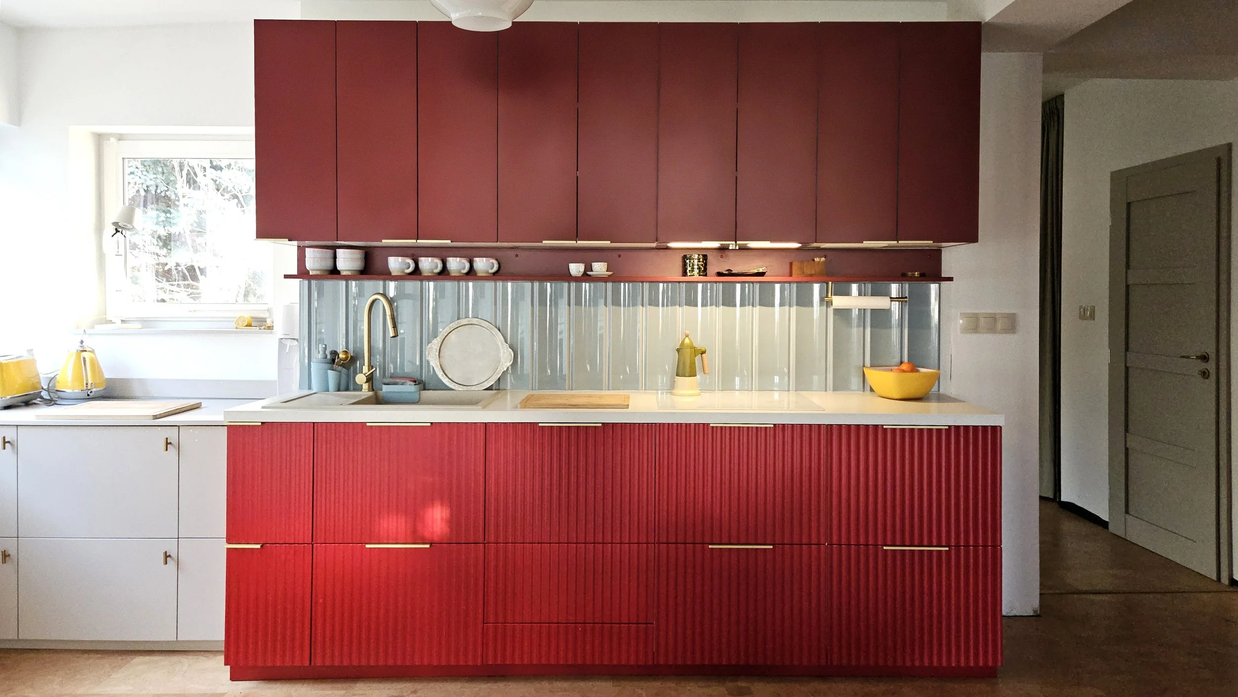 Modern kitchen with red cabinets, a white countertop, gold accents, a window with a view of trees, and yellow kitchen appliances.