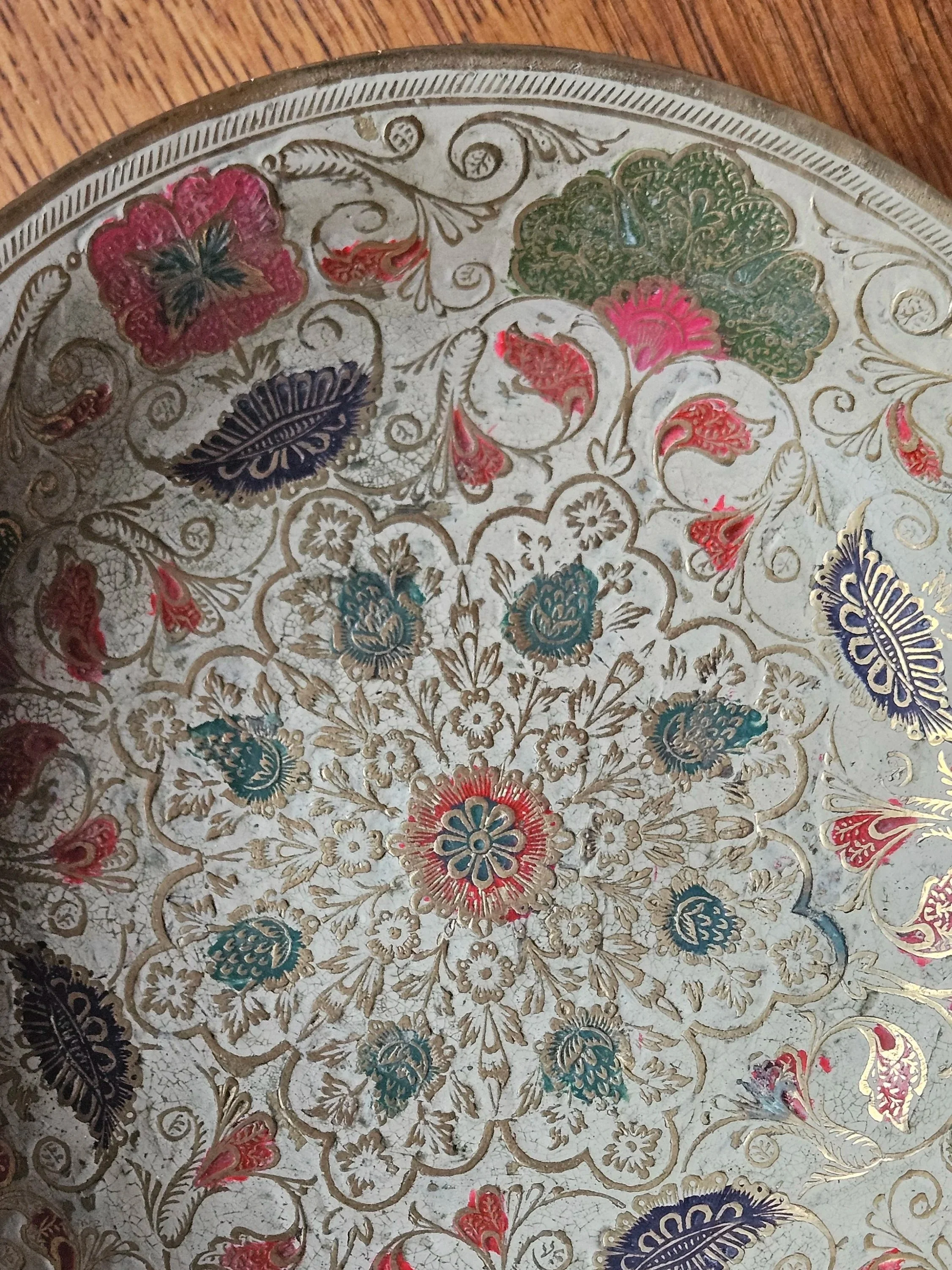 Decorative metal tray with intricate floral and leaf patterns in red, blue, green, and gold, on a wooden surface.