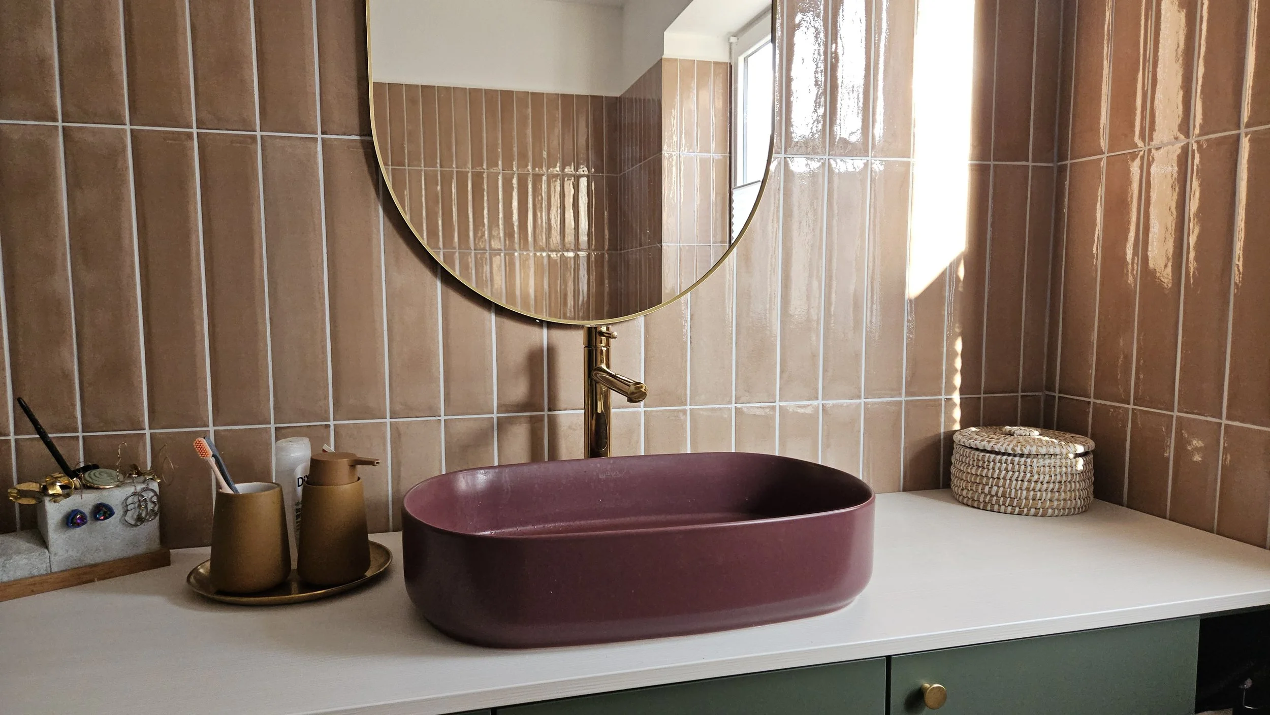 A bathroom vanity with a maroon countertop basin, a rectangular mirror, a gold faucet, and various bathroom accessories, with tan tiled walls and a window letting in sunlight.