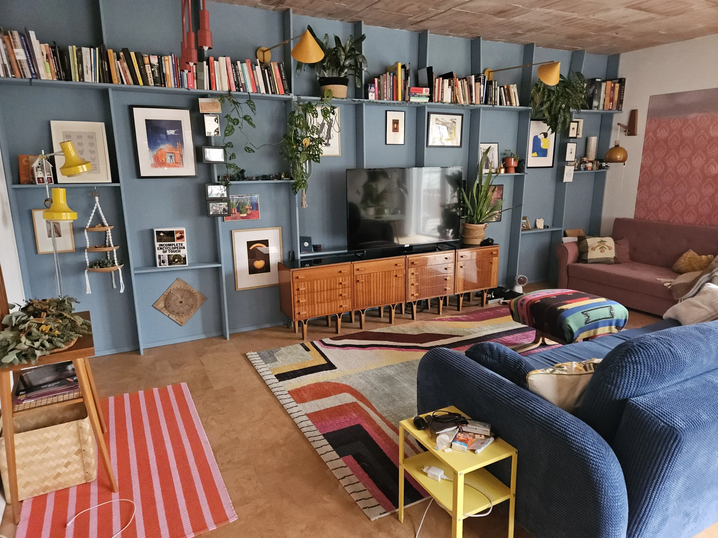 A cozy living room with a blue wall featuring a built-in bookshelf, framed art, plants, and decorative items. There are colorful area rugs, couches, a yellow side table with items on it, and a wooden TV stand with a large flat-screen TV.