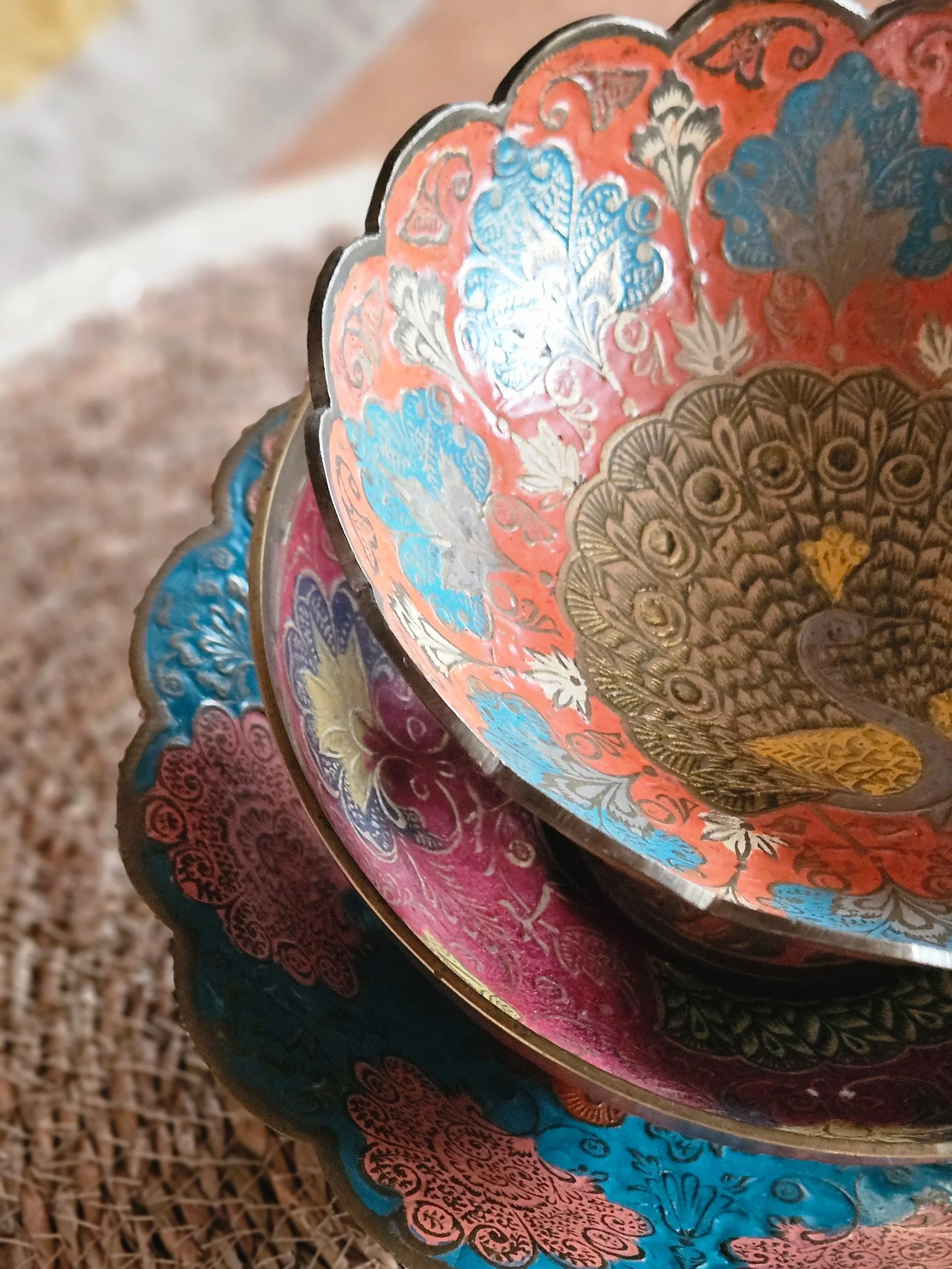 Close-up of decorated metal bowls with floral patterns in orange, blue, pink, and gold, stacked on a woven surface.