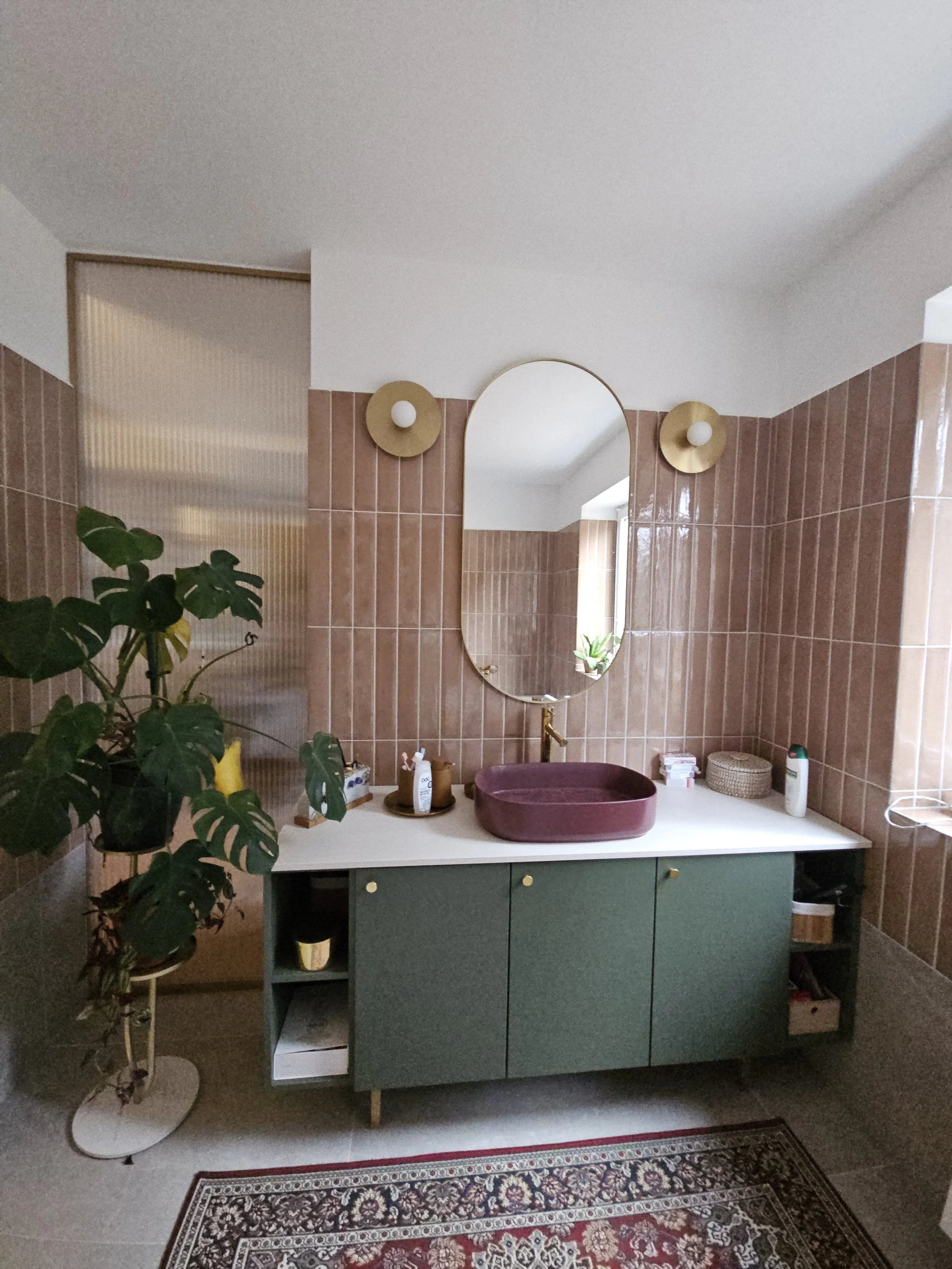 A bathroom vanity with a green cabinet, white countertop, and a maroon vessel sink, with a large oval mirror above and two wall-mounted lights, a plant on the left, and various toiletries and decor items on the counter.