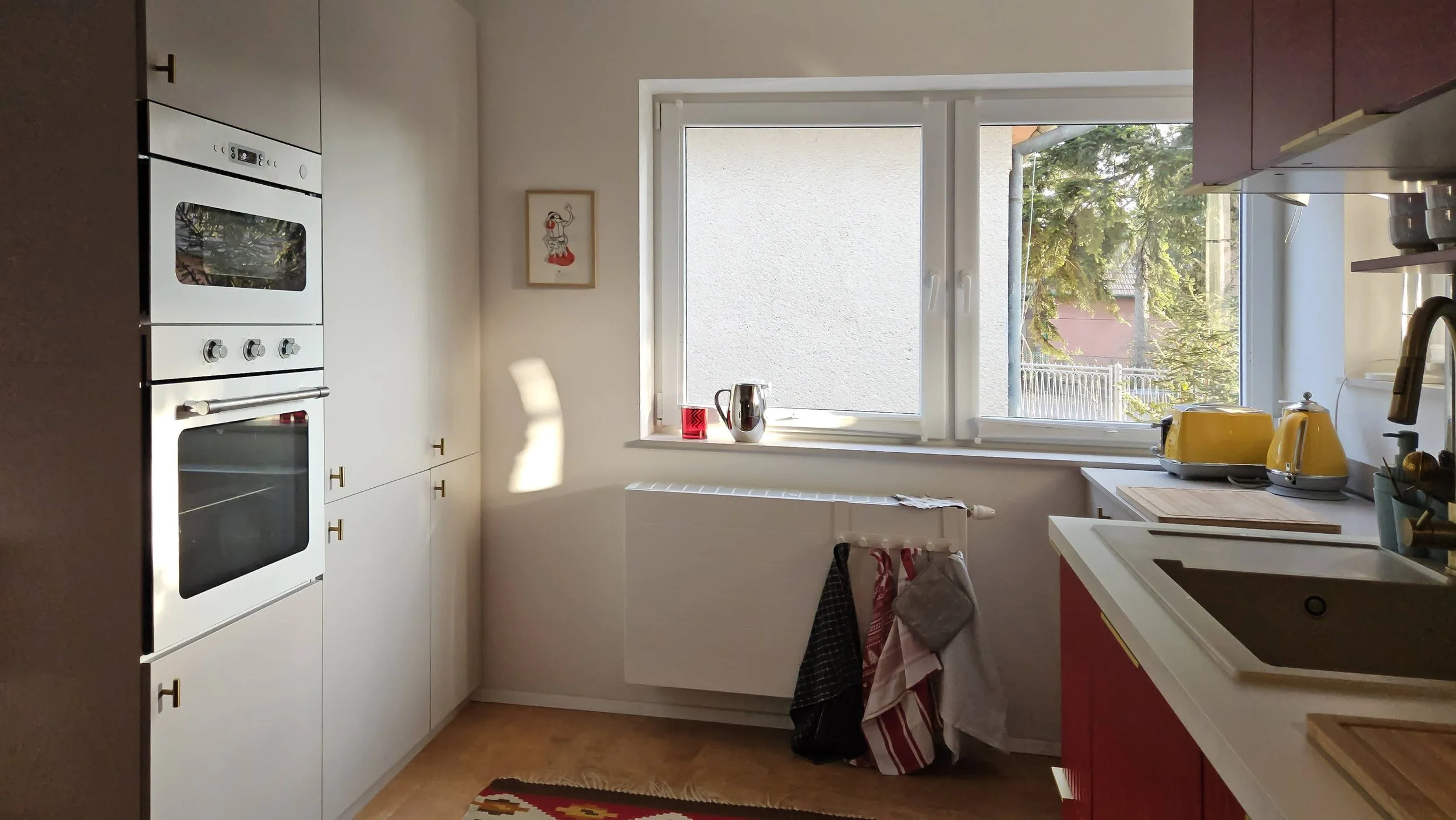Kitchen with white cabinets, a window with a view of trees, yellow toaster and kettle on the counter, and a red cabinet under the sink.
