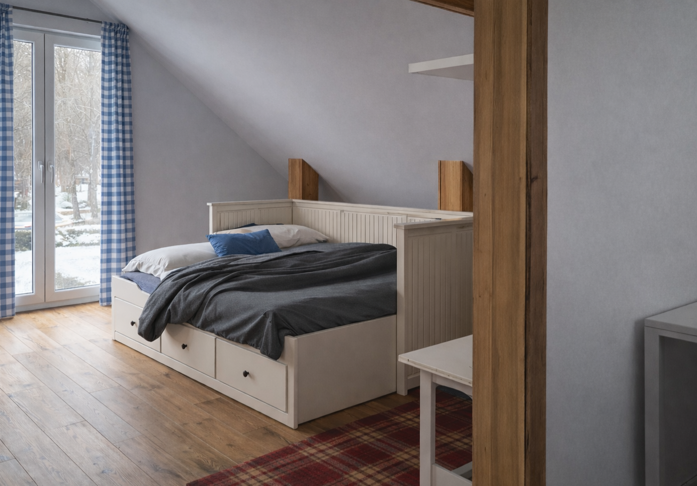 A cozy bedroom with a white bed featuring storage drawers, against a sloped ceiling wall with wooden beams, next to glass doors with blue checkered curtains showing a snowy outdoor scene.