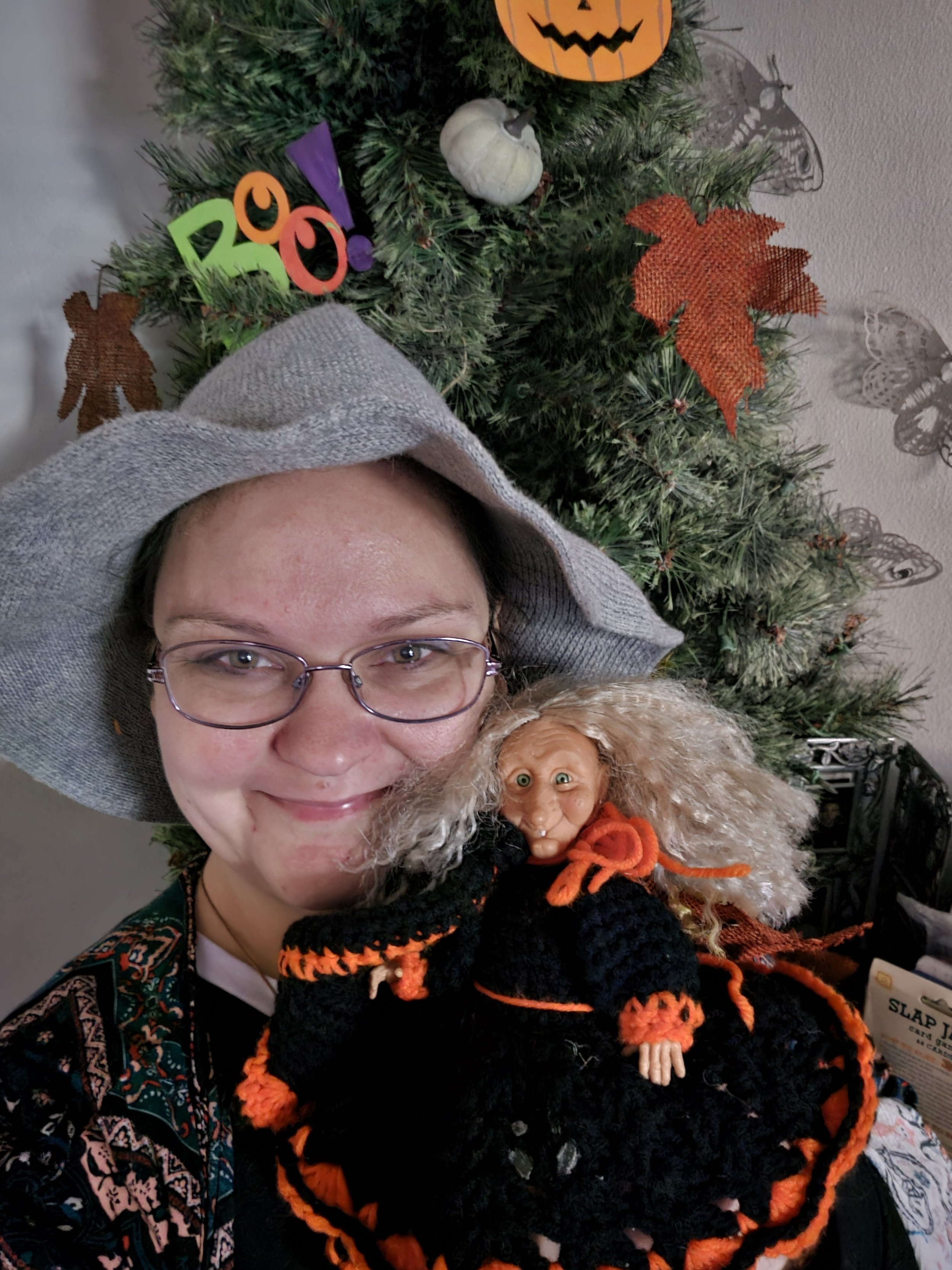 A smiling woman with glasses and a gray headscarf holds a Halloween doll in front of a decorated Christmas tree. The tree has orange and black themed ornaments, including a pumpkin and fall leaves.