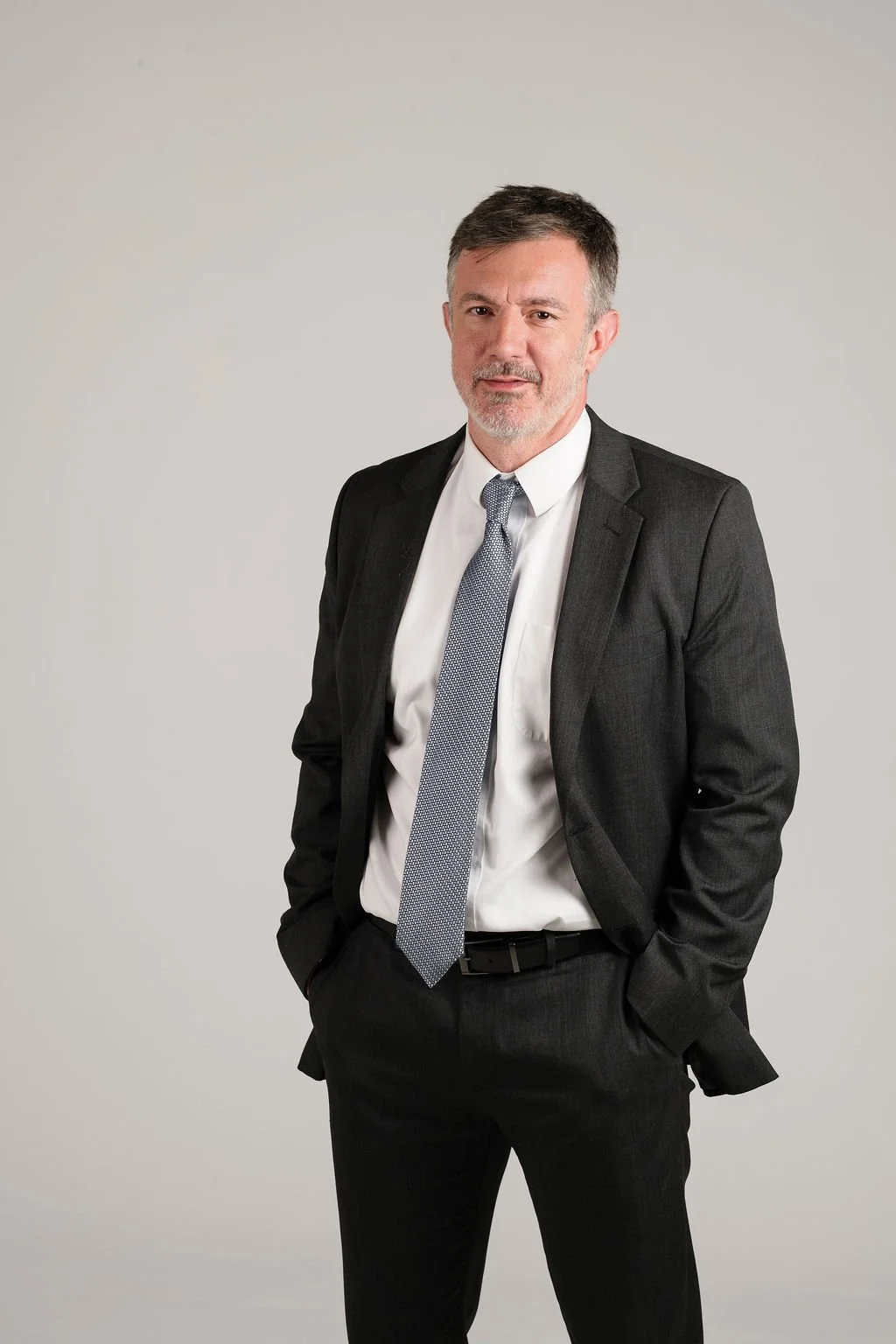 A middle-aged man in a black suit, white dress shirt, and gray patterned tie stands against a plain light gray background.