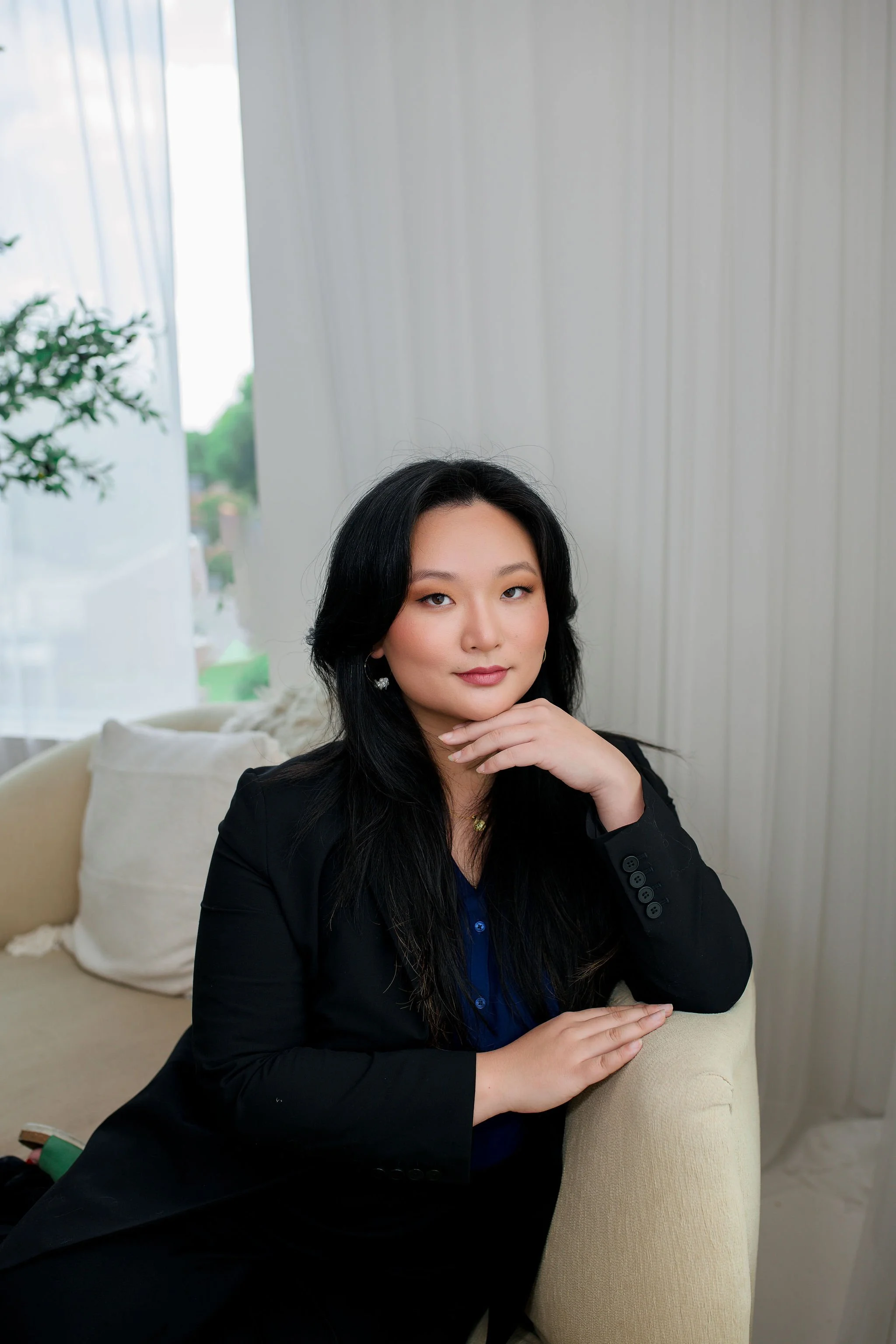A woman with long black hair and a black blazer sitting on a light-colored sofa in a well-lit room with white walls and sheer curtains, looking at the camera with a confident expression.