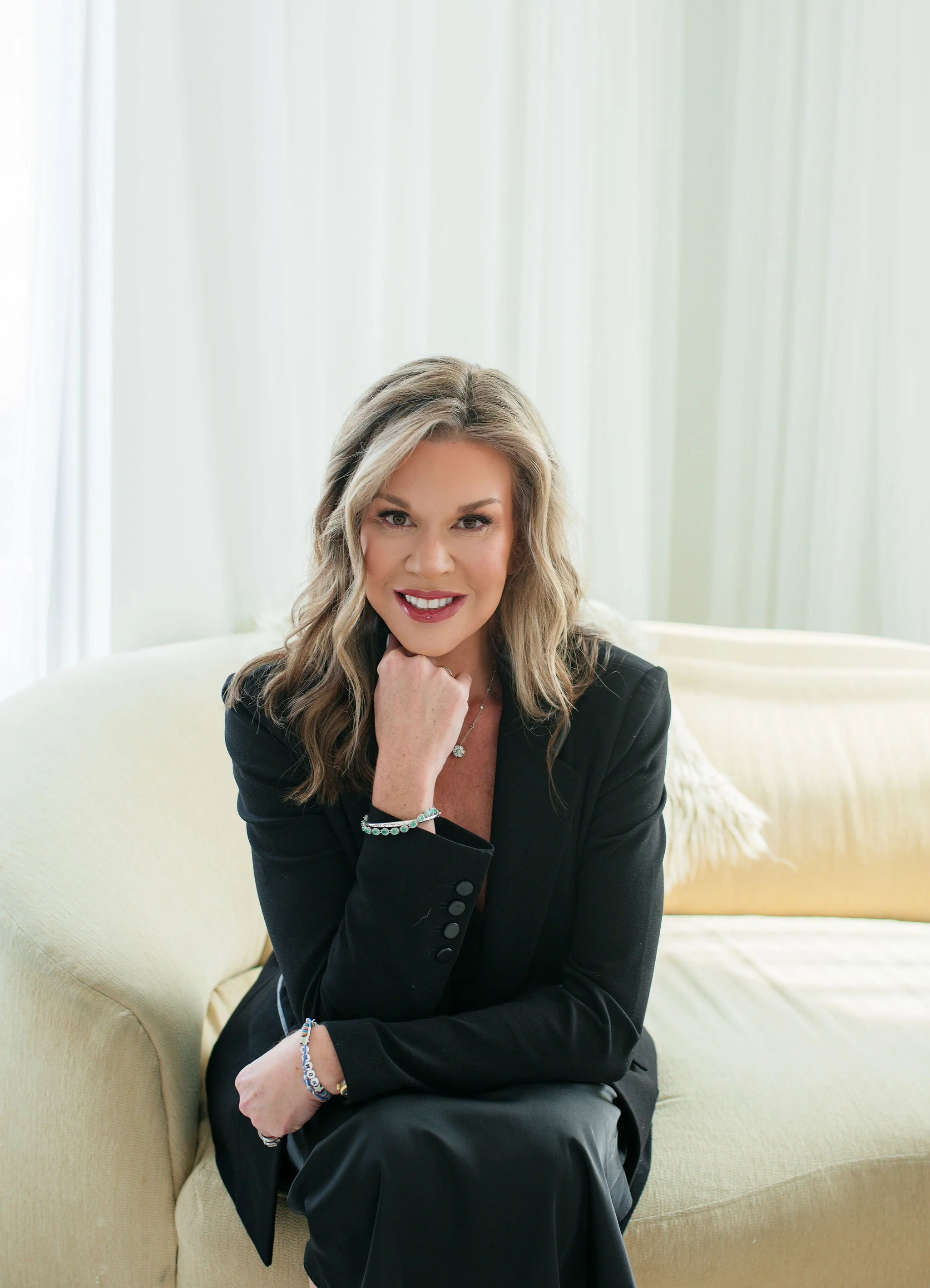 A woman with blonde hair wearing a black blazer and sitting on a light-colored sofa, smiling with her hand on her chin, in a bright room with white curtains.