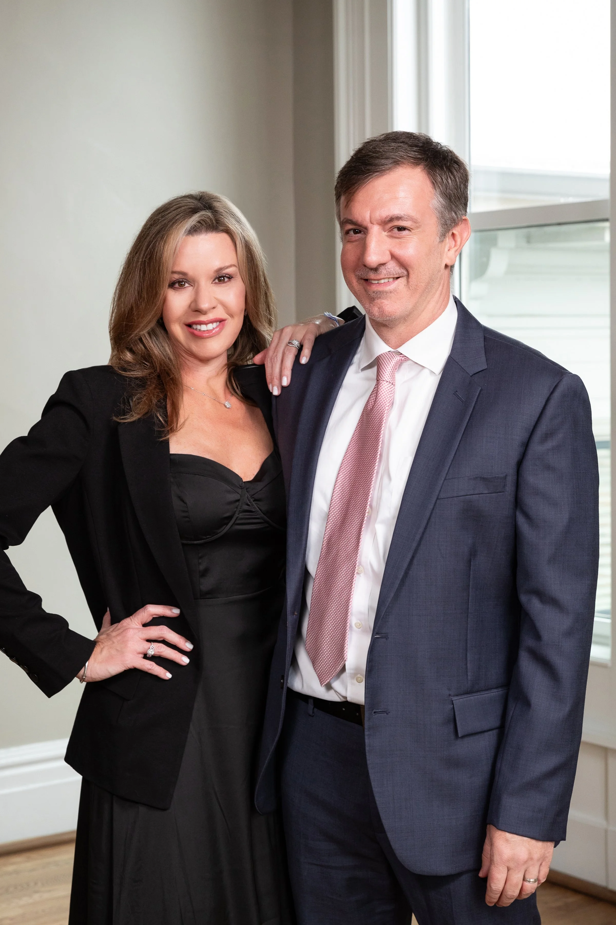 A man and woman dressed in formal attire posing together indoors near a window, smiling at the camera.