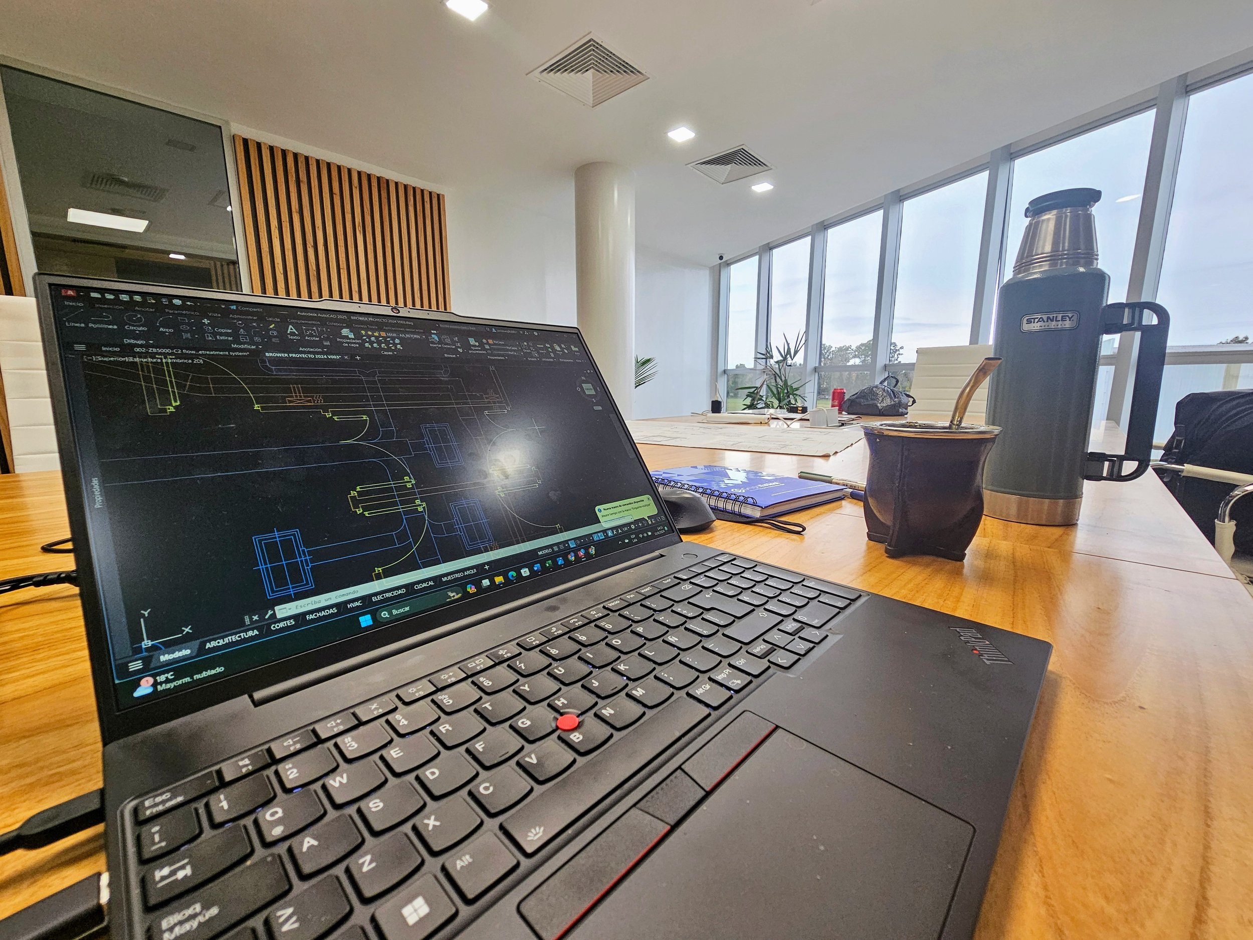 Una mesa de oficina con una laptop encendida mostrando un diseño técnico, junto a una botella térmica, un vaso con una bebida y otros objetos en un espacio con ventanas grandes y luz natural.