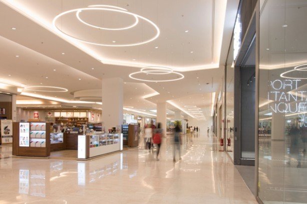 Interior de un centro comercial moderno con iluminación circular en el techo y tiendas a la derecha.