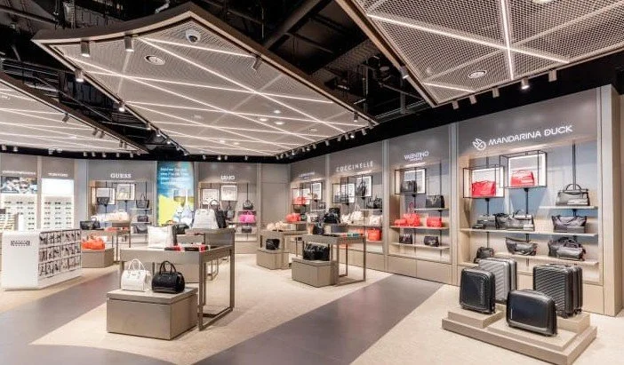 Interior de una tienda de bolsos y accesorios de marca Mandarina Duck con exhibidores de bolsas, maletas y carteras en colores diversos.