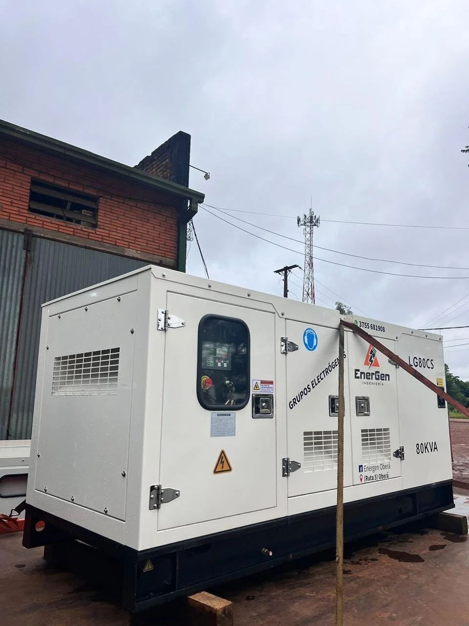 Generador eléctrico en exteriores sobre superficie de concreto, con cables y edificios en el fondo, cielo nublado.